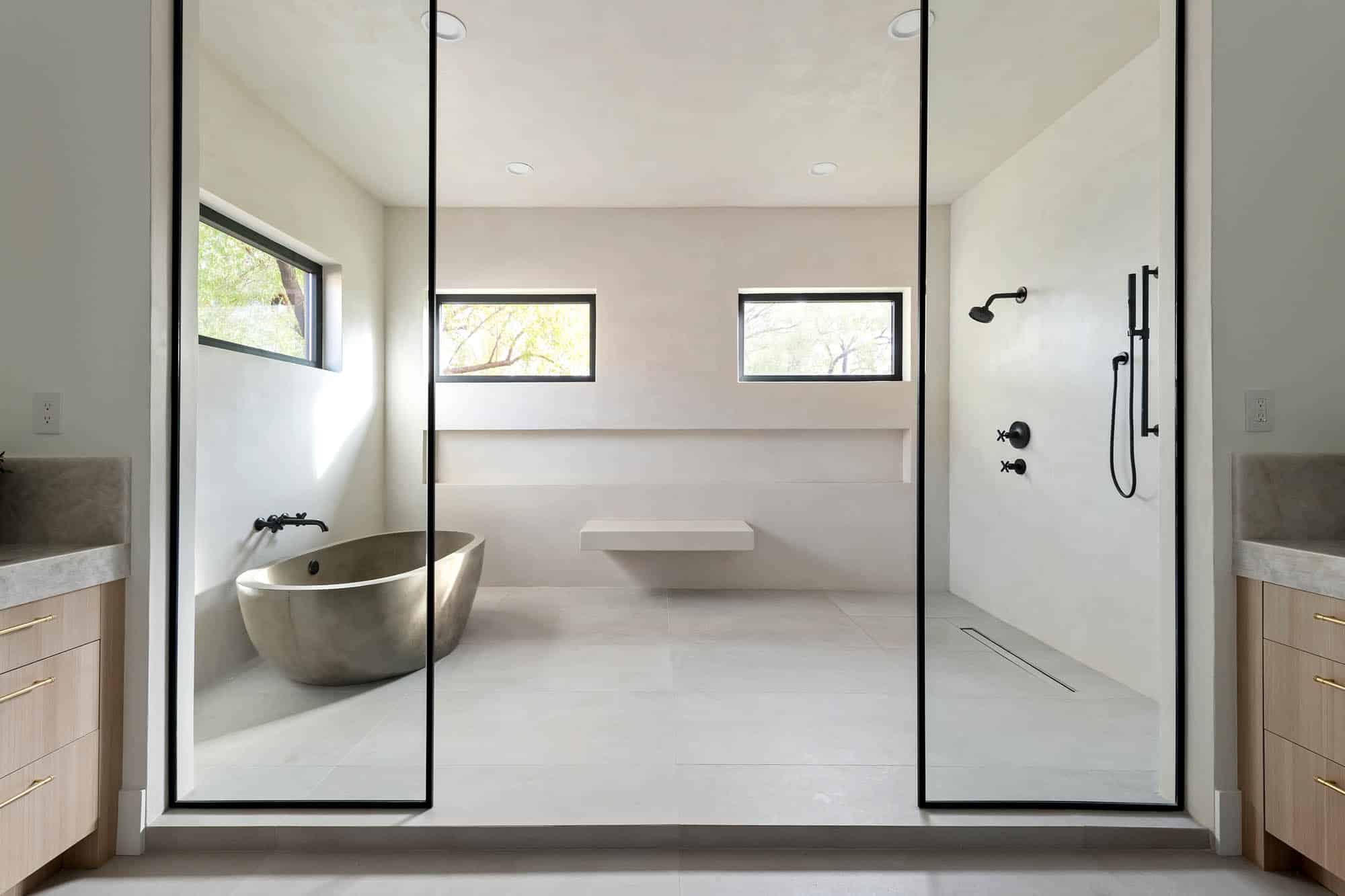 Primary bathroom wet room behind floor-to-ceiling glass enclosure with black frames, freestanding concrete soaking tub with matte black wall-mounted filler, floating bench, walk-in shower with matte black shower head and handheld sprayer, three clerestory windows with tree views, linear drain, and light wood vanity cabinetry visible on both sides at The Carmella by Kingdom & Co.