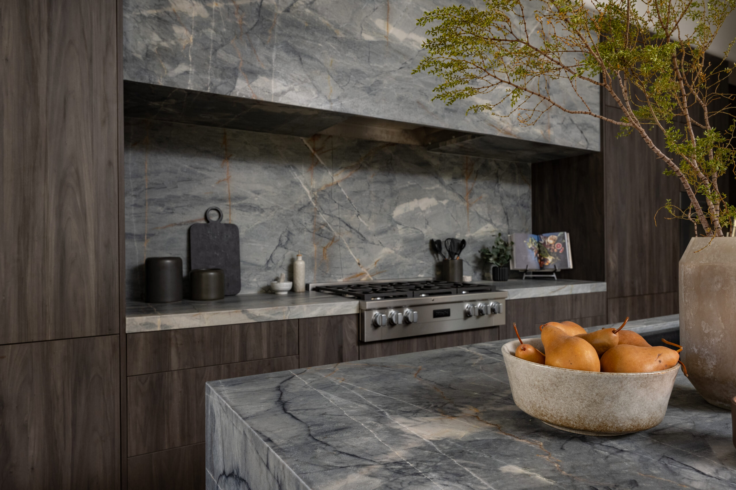 Contemporary Marble Kitchen Design Modern kitchen with a sleek marble countertop and backsplash, featuring a gas stove, dark wood cabinets, and a bowl of pears. Decorated with potted plants, a cutting board, and kitchen utensils, creating a stylish and functional cooking space.