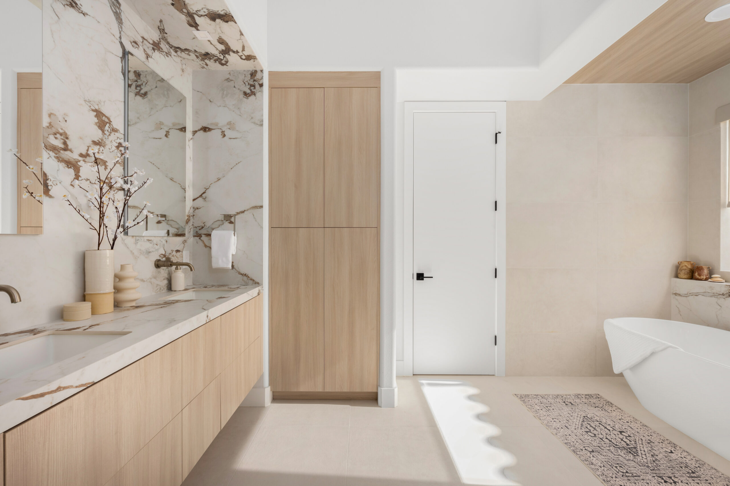 Primary bathroom wide-angle view with light oak floating vanity, marble countertop, white and brown marble accent wall, frameless LED-backlit mirror, tall oak linen cabinet, white door, and freestanding soaking tub visible in adjacent alcove in The Manhattan Lake Las Vegas remodel by Kingdom & Co.