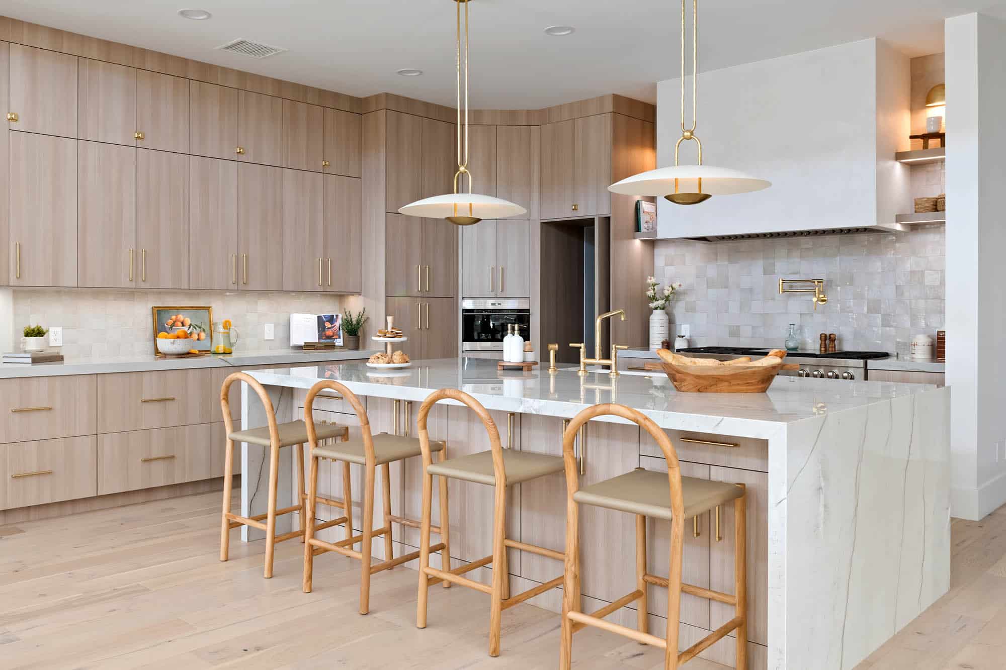 Kitchen with light wood flat-panel cabinetry, marble waterfall island with rattan bar stools, brass pendant lights, brass hardware and faucet, zellige tile backsplash, floating wood shelves, professional range with custom hood at The Carmella by Kingdom & Co.
