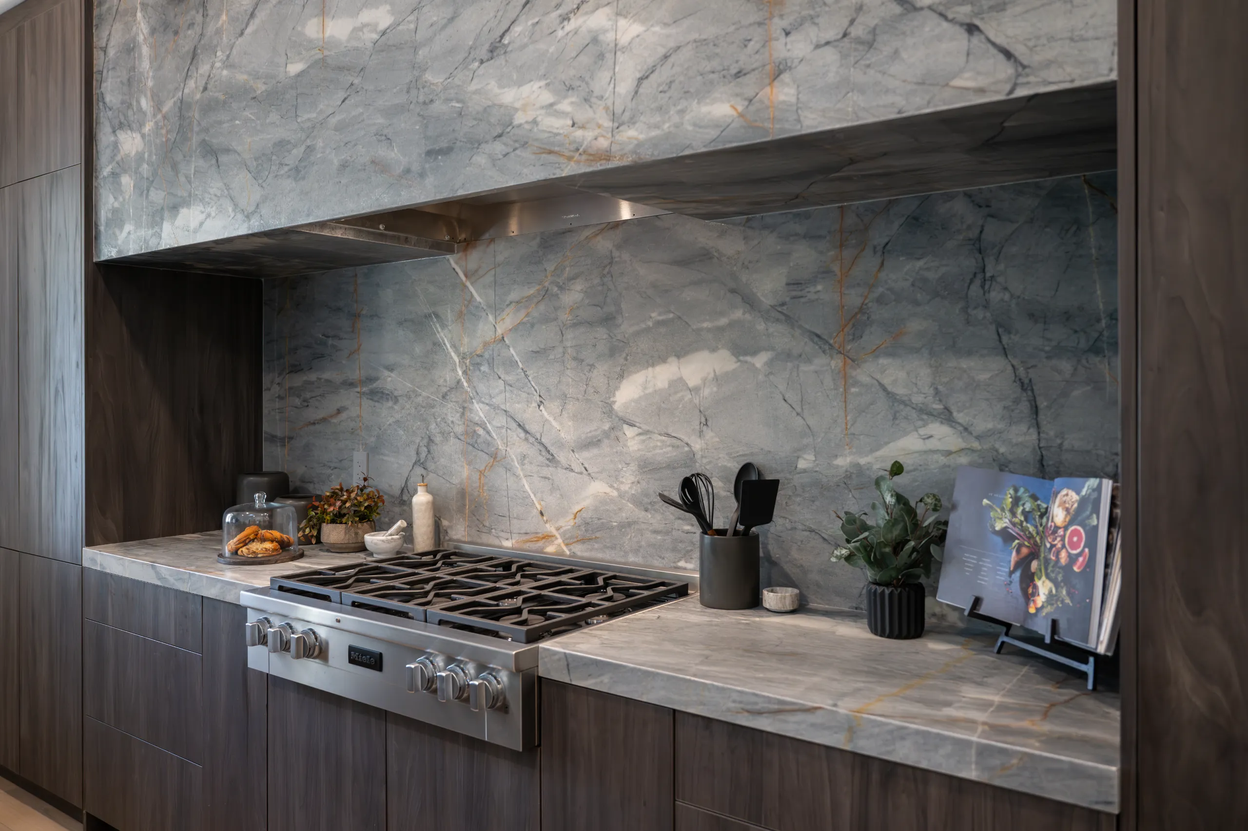 Luxurious Marble Kitchen Stove Design Modern kitchen with a stunning marble backsplash and countertop, featuring a gas stove, dark wood cabinets, and decorative elements like candles, potted plants, utensils, and a cookbook stand, creating a luxurious and functional cooking area.