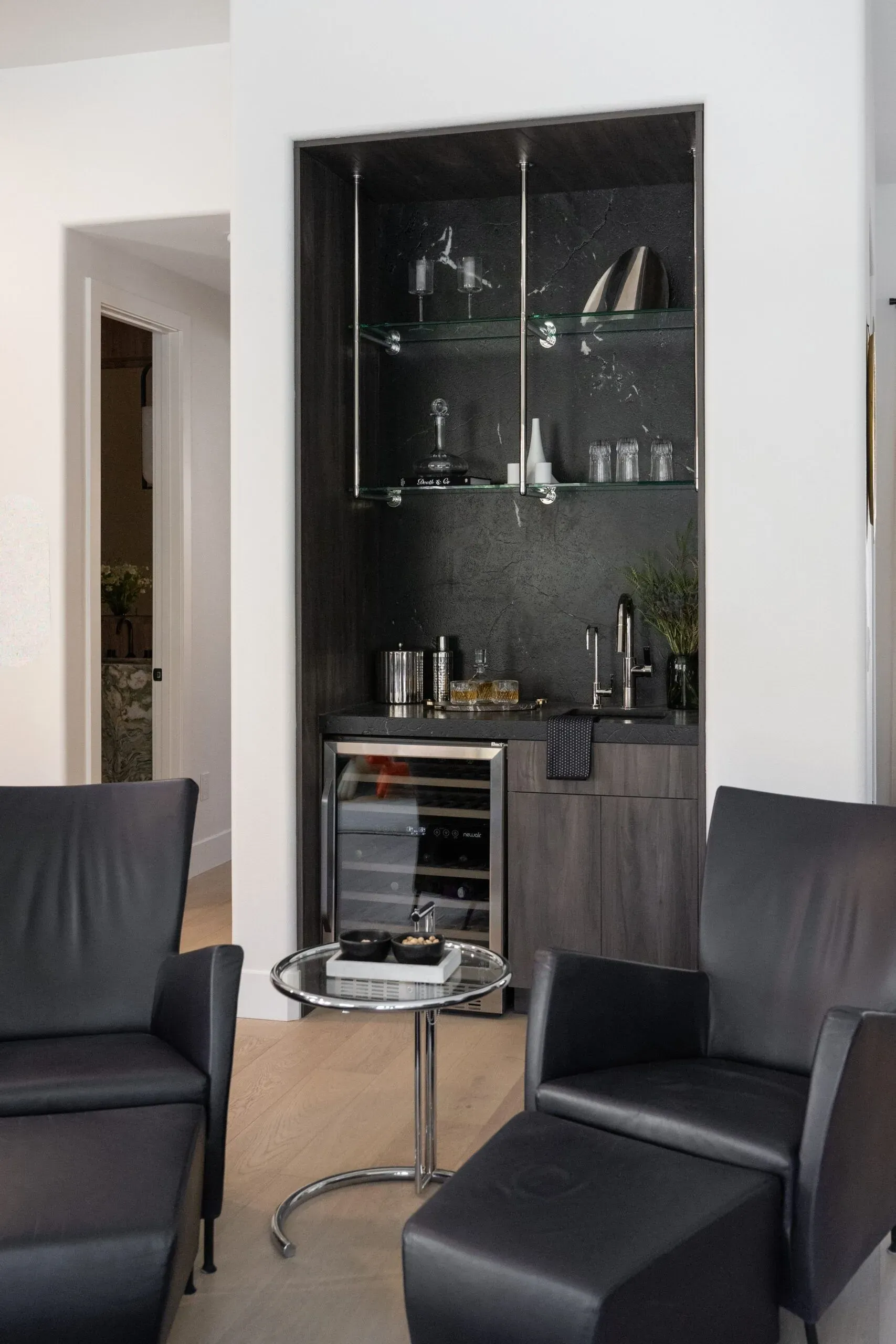Chic Modern Home Bar Design Modern home bar area with a sleek black cabinet, glass shelves displaying barware, and a built-in wine cooler. Adjacent to black leather chairs and a glass side table, creating a stylish and functional space for entertaining.