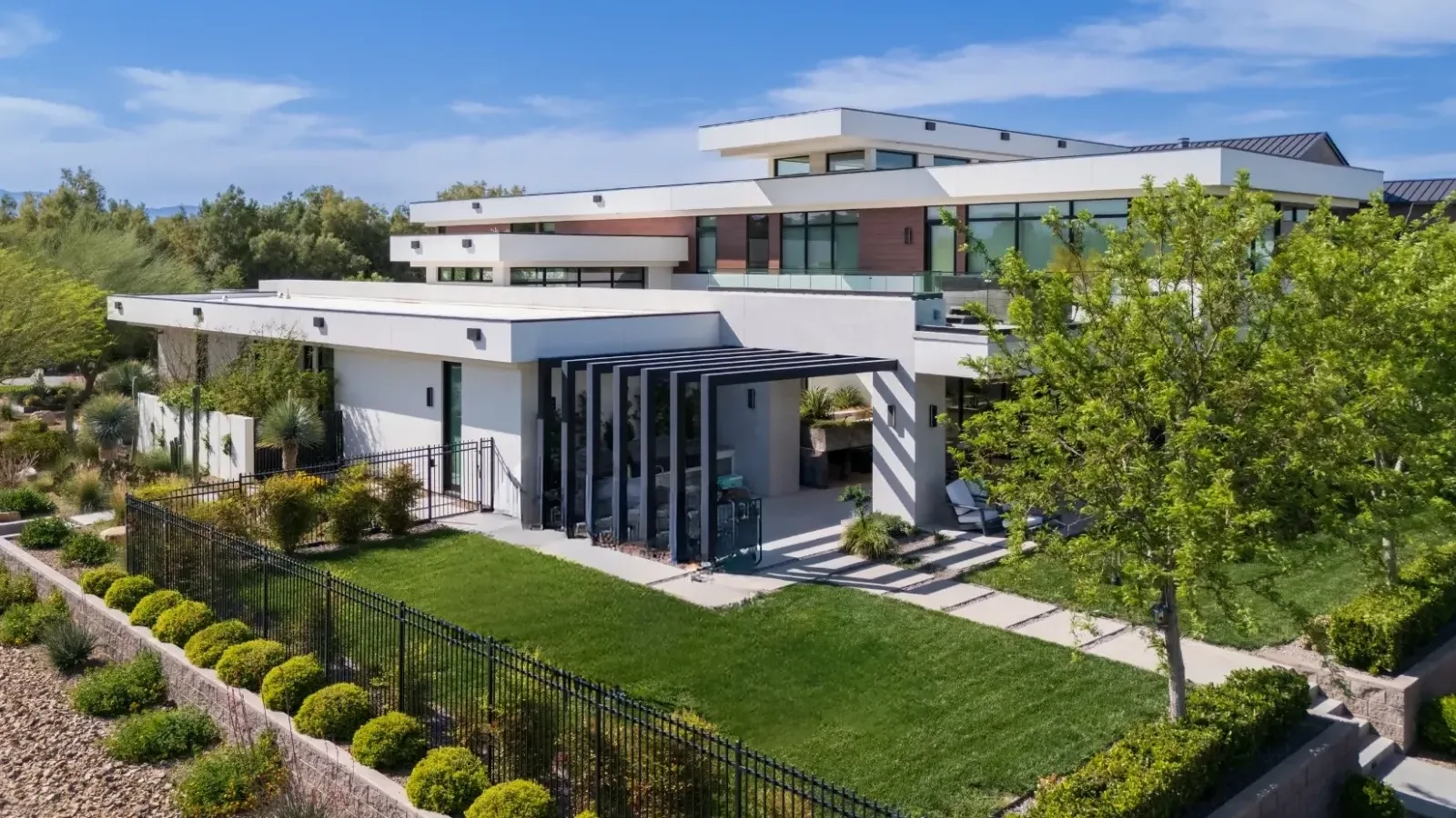 Custom luxury new build by Kingdom & Co in Las Vegas, featuring a modern two-story design with white exterior walls, large glass windows, and a flat roof. Surrounded by lush green landscaping and a well-maintained lawn, ideal for upscale desert living.