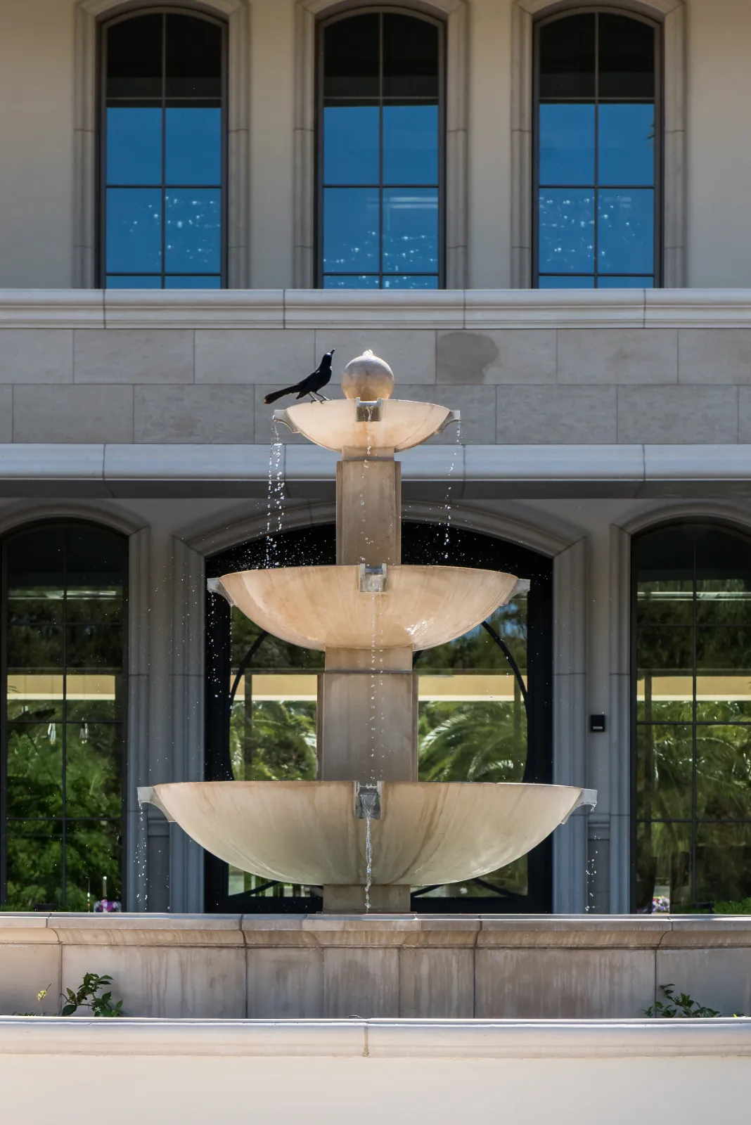 English Estate – Elegant Multi-Tiered Fountain by Kingdom & Co in Las Vegas Close-up of a multi-tiered fountain at English Estate, a luxury home by Kingdom & Co in Southern Highlands, Las Vegas, with cascading water, stone construction, and a backdrop of arched windows on a beige stone facade, featuring a bird perched on top.