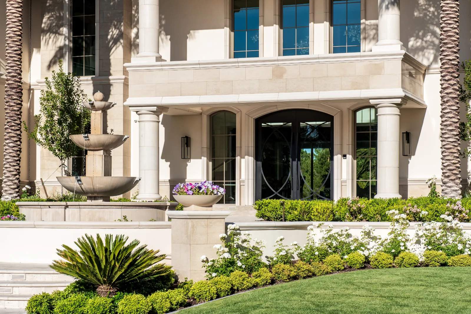 English Estate – Grand Entrance with Fountain by Kingdom & Co in Las Vegas Close-up of the grand entrance at English Estate, a luxury Mediterranean-style home by Kingdom & Co in Southern Highlands, Las Vegas, featuring a beige stone facade, tall columns, large arched windows, a multi-tiered stone fountain, green shrubs, palm trees, and lush landscaping.
