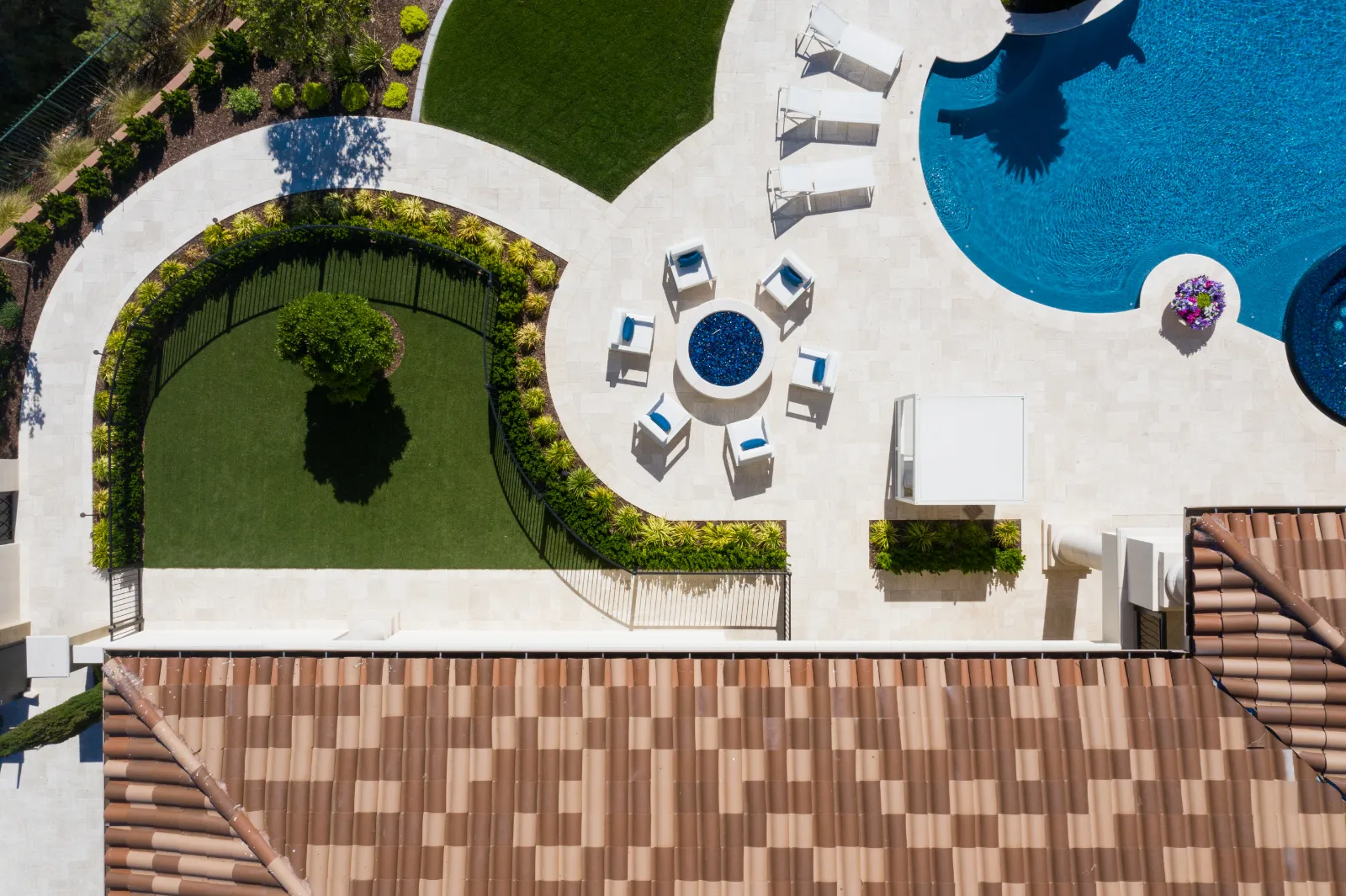 English Estate – Luxury Outdoor Area with Fire Pit and Pool by Kingdom & Co in Las Vegas Aerial view of English Estate's luxury outdoor area by Kingdom & Co in Southern Highlands, Las Vegas, featuring a red-tiled roof, circular fire pit with white seating, freeform blue pool, green lawns, and curved landscaping with shrubs.
