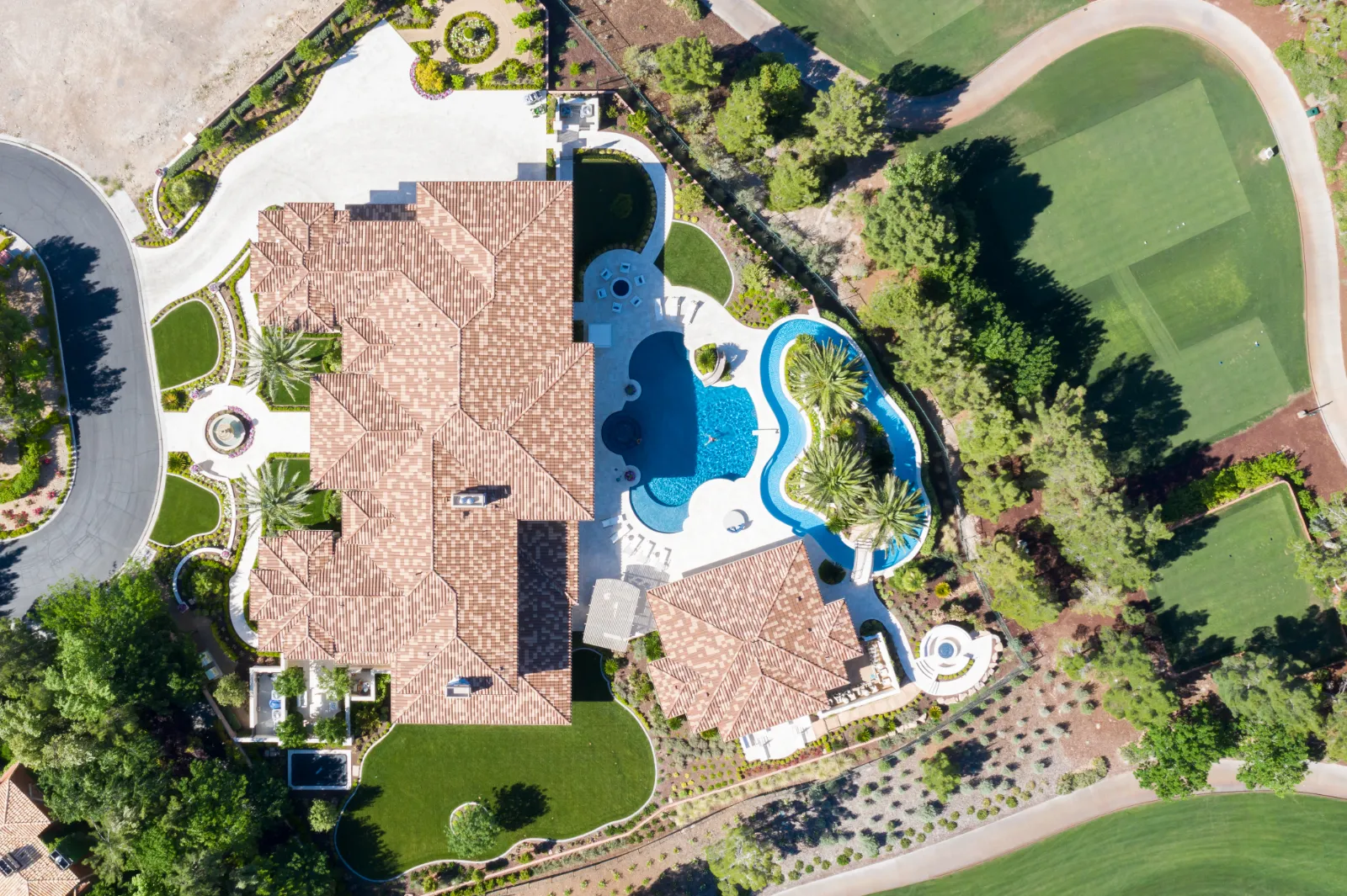 English Estate – Aerial View of Luxury Home with Pool by Kingdom & Co in Las Vegas Aerial view of English Estate, a luxury Mediterranean-style home by Kingdom & Co in Southern Highlands, Las Vegas, featuring a red-tiled roof, freeform blue pool, green lawns, palm trees, and surrounding desert landscaping with winding pathways.