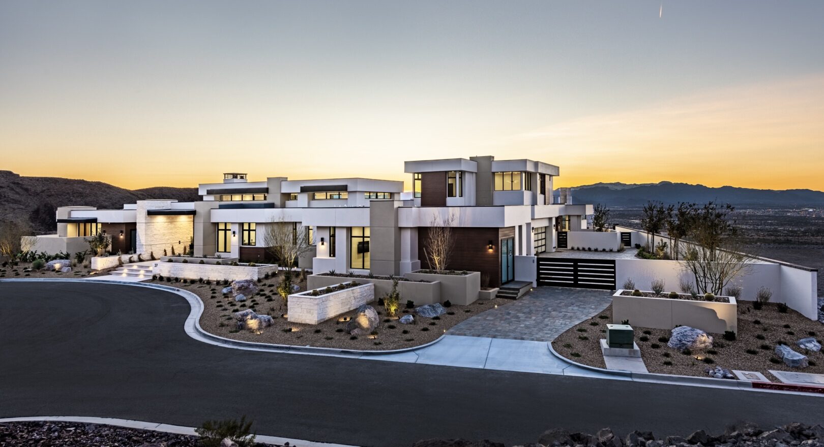 Luxurious modern custom home by Kingdom & Co, featuring a multi-level design with white and wood accents, large windows, and a curved driveway. Set against a stunning sunset backdrop with desert landscaping and a gated entrance, ideal for upscale living.