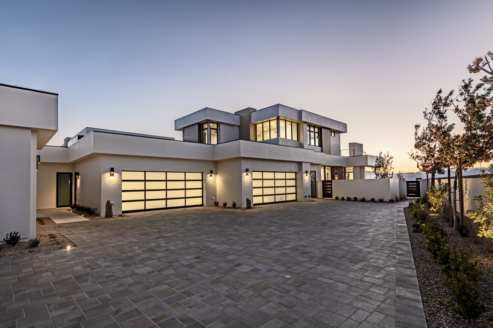 Luxurious modern custom home built by Kingdom & Co, featuring a sleek two-story design with large glass garage doors, white exterior walls, and a spacious paver driveway. Surrounded by landscaped greenery and illuminated by stylish outdoor lighting, ideal for upscale living.