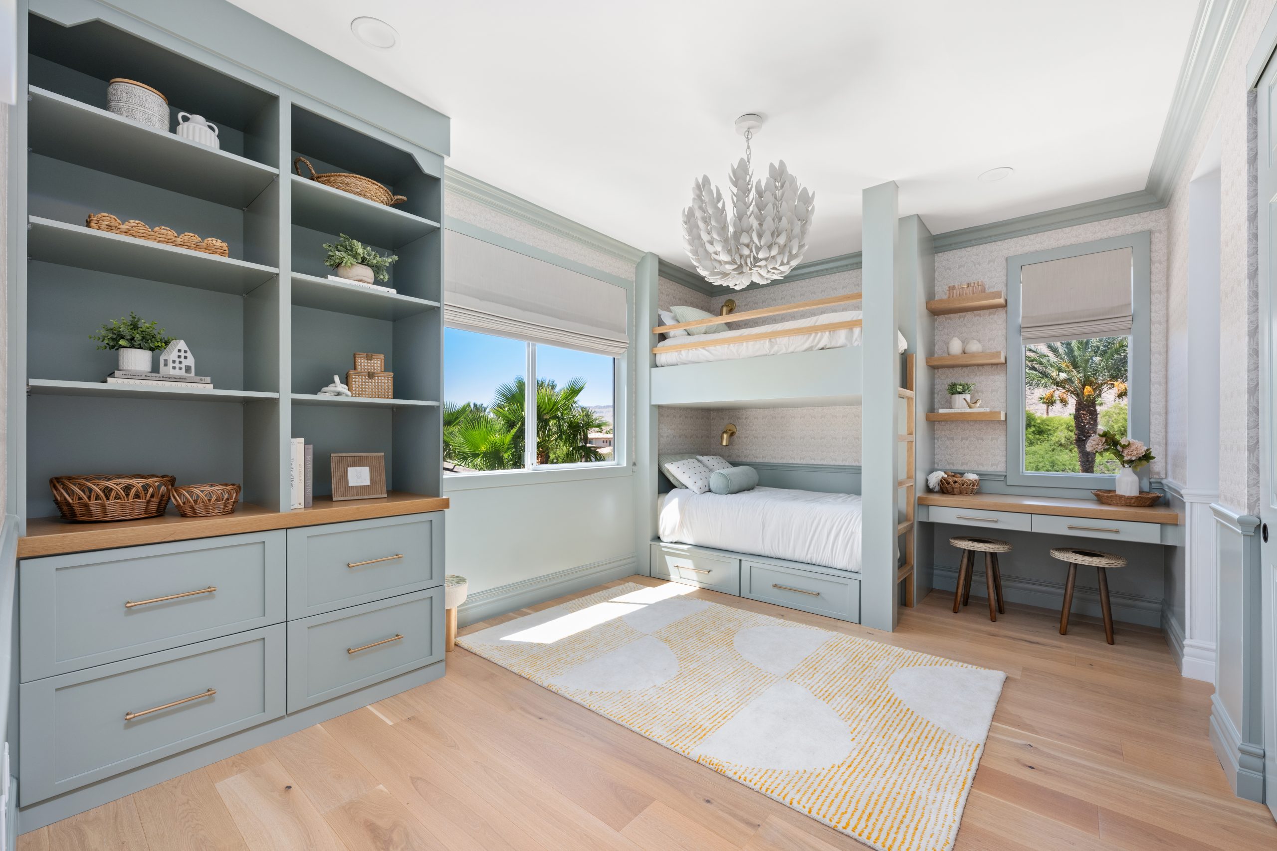 Kids bunk room with built-in blue-gray bunk beds, natural wood ladder and accents, custom built-in shelving with brass hardware and wood countertop, white leaf chandelier, patterned wallpaper, and yellow polka dot area rug at The Winchester home remodel by Kingdom & Co.