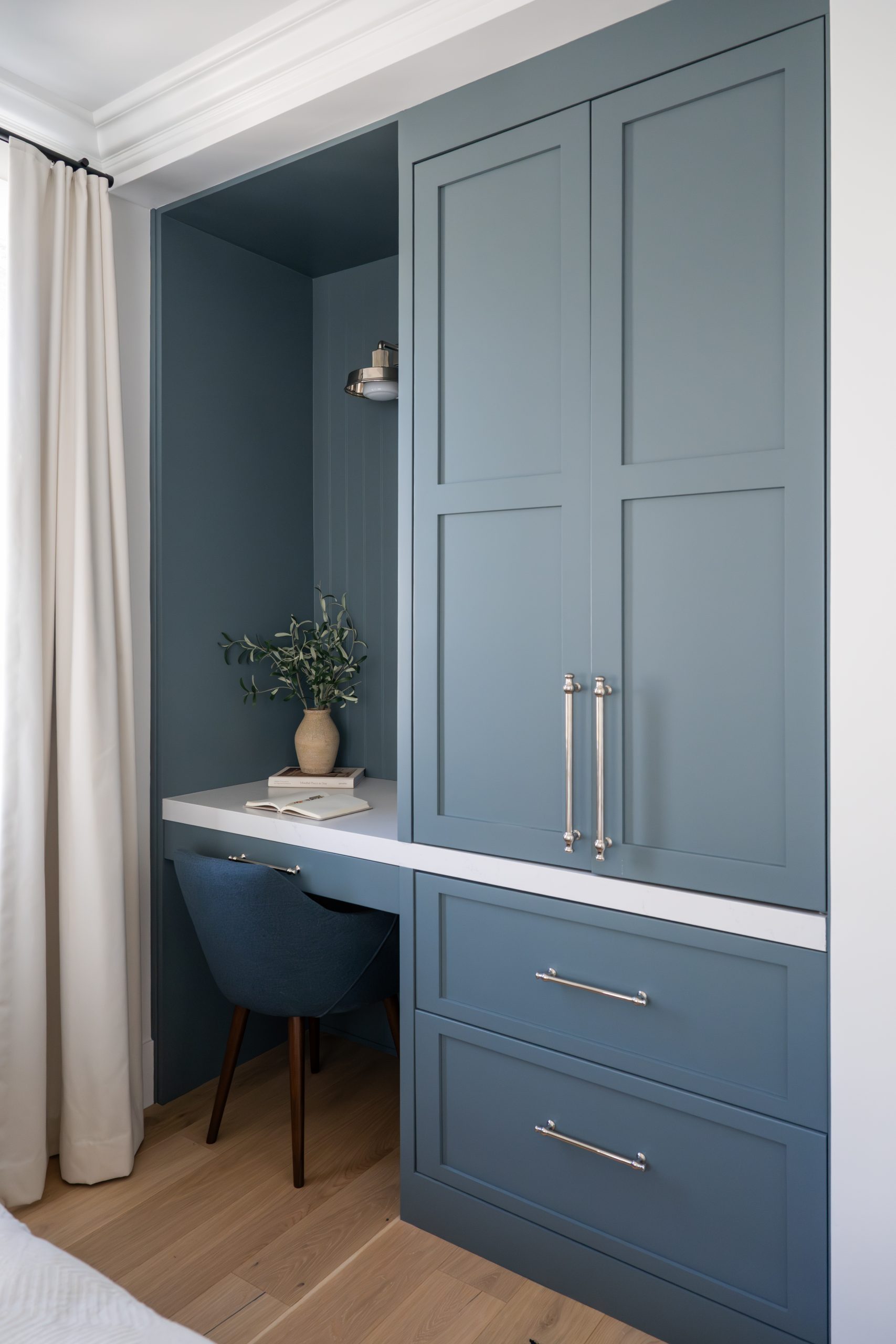 Built-in desk nook with blue-gray cabinetry, white quartz countertop, polished nickel hardware, brass wall sconce, blue upholstered chair, beadboard paneling, and styled vase with greenery at The Winchester home remodel by Kingdom & Co.