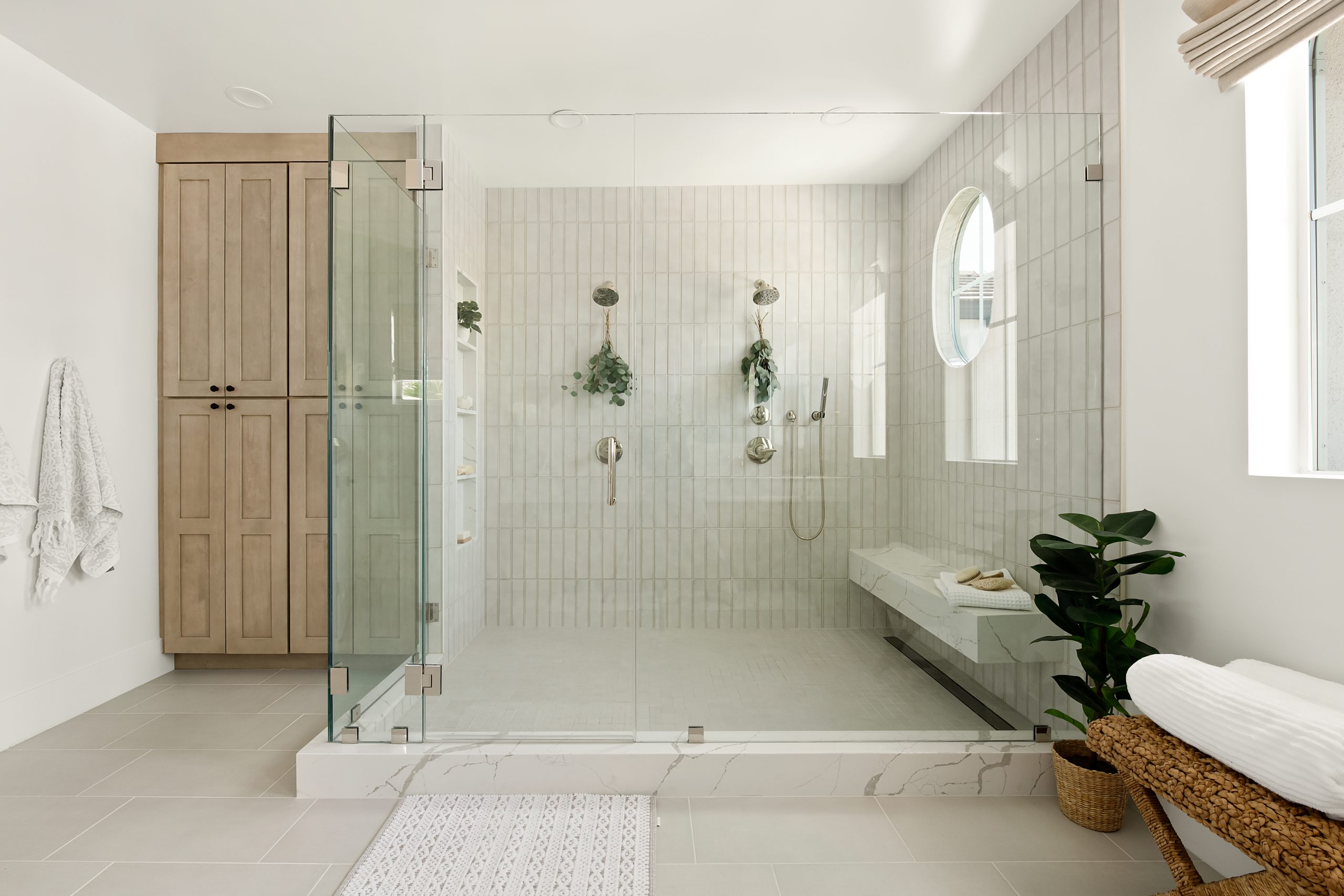Primary bathroom with large glass-enclosed walk-in shower, vertical stacked tile walls, built-in marble shower bench, oval window, brushed nickel fixtures, light wood linen cabinet, and woven rattan bench with potted plant at The Winchester home remodel by Kingdom & Co.
