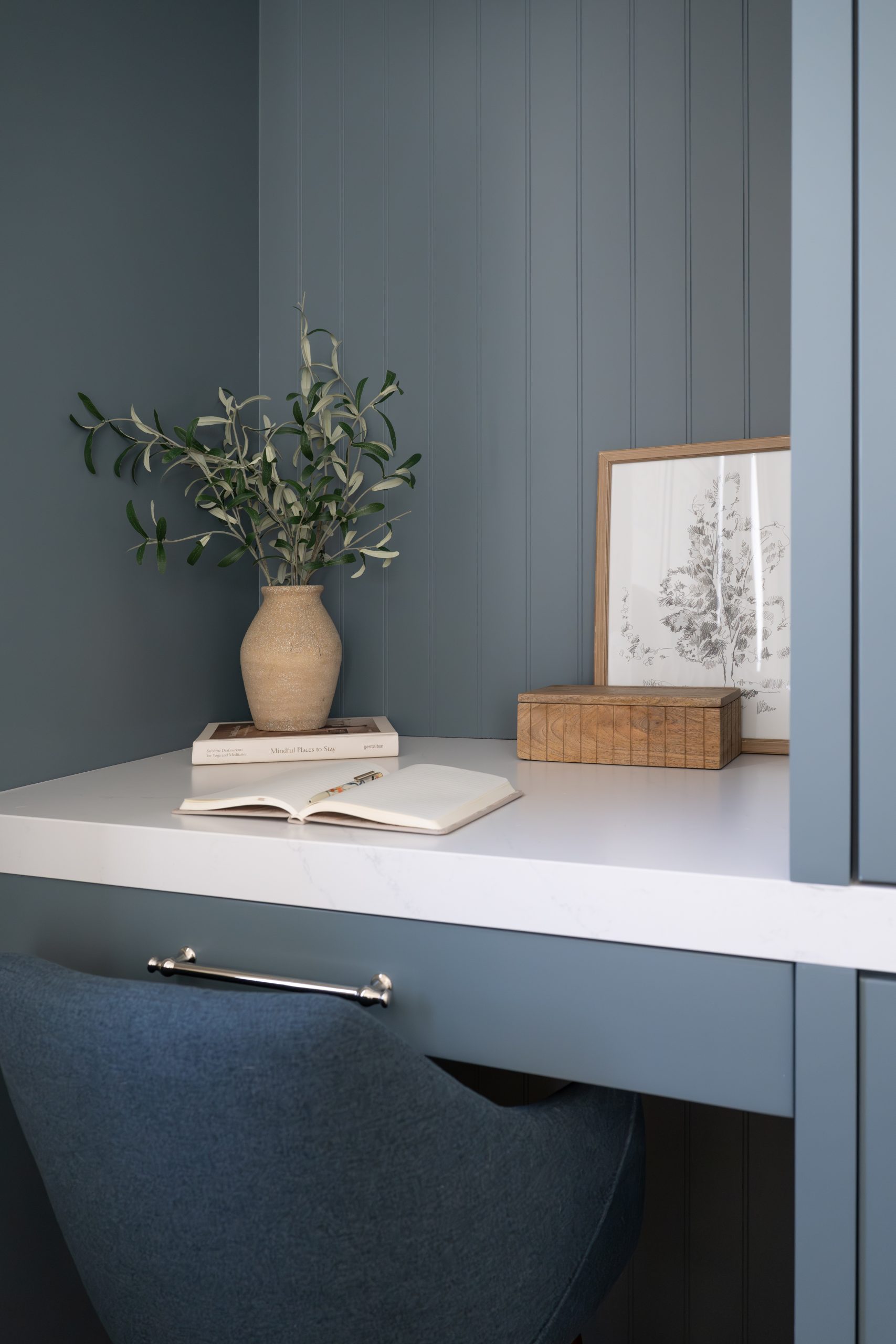 Close-up of built-in desk nook detail with blue-gray beadboard paneling, white quartz desktop, open book, ceramic vase with greenery, framed tree sketch, and wood decorative box at The Winchester by Kingdom & Co.