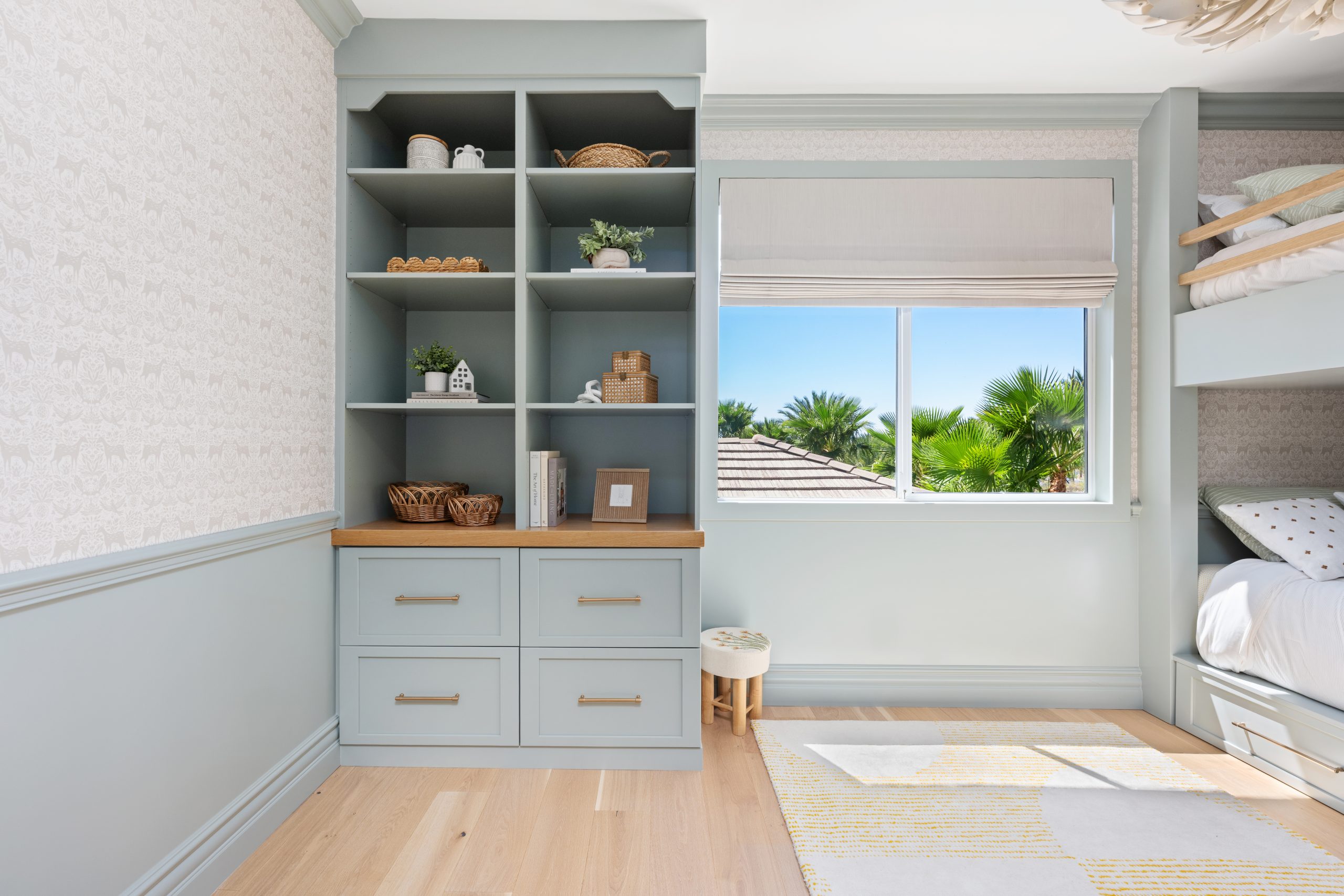 Kids bunk room alternate view with blue-gray built-in shelving, natural wood countertop, brass hardware, window with Roman shade and palm tree views, patterned wallpaper, and built-in bunk beds at The Winchester home remodel by Kingdom & Co.
