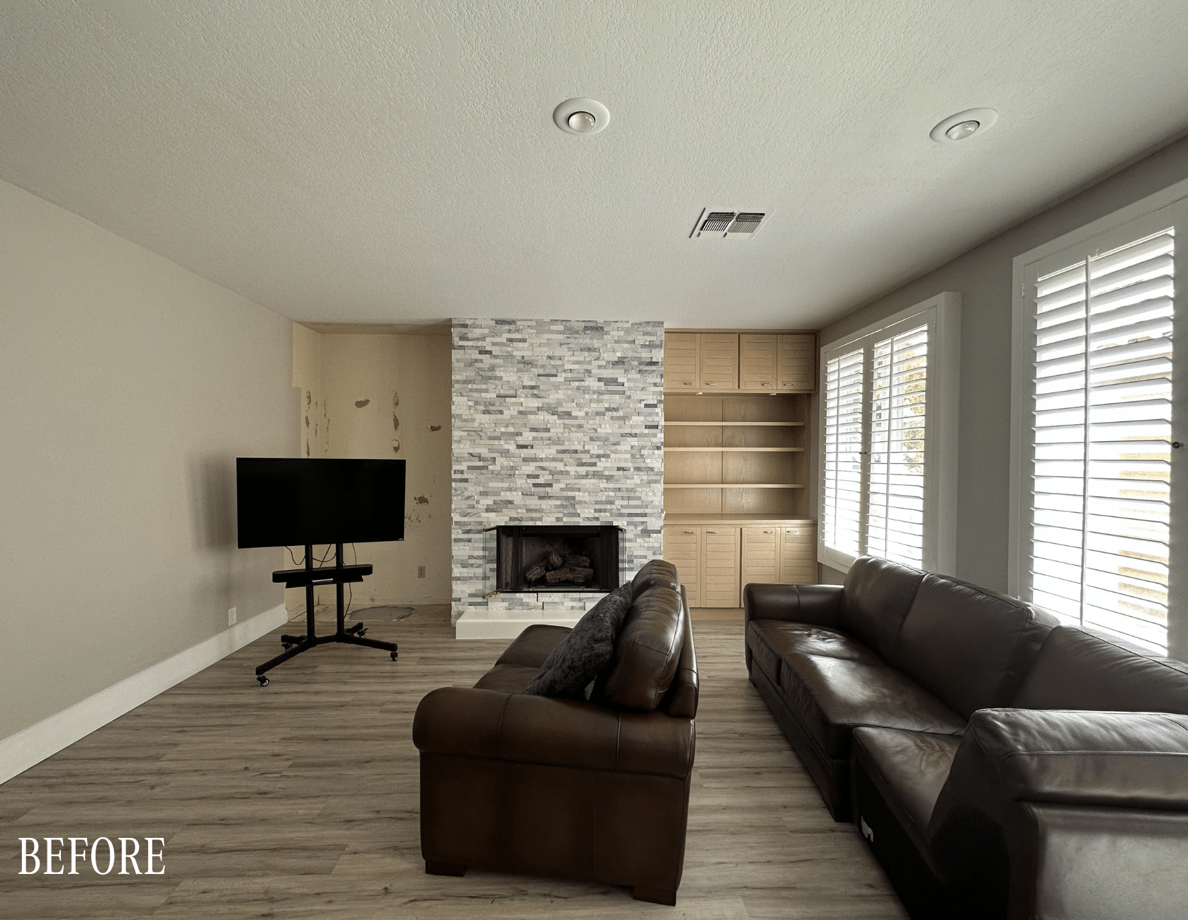 Before: Original living room with brick corner fireplace, brown leather furniture, and shuttered windows in The Parker