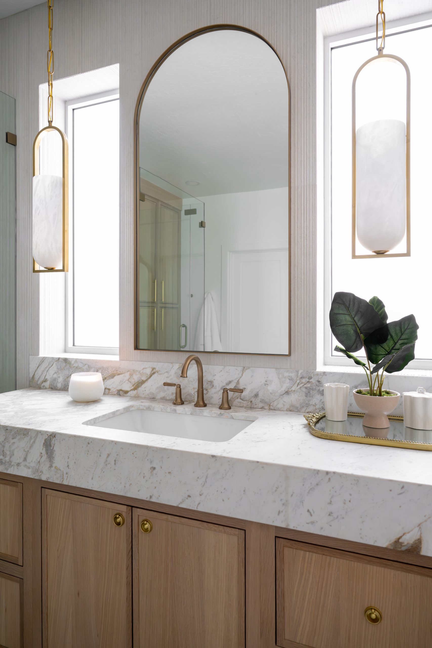 Single vanity with arched brass mirror, alabaster and brass pendant lights, white oak shaker cabinetry, and calacatta marble waterfall countertop at The Charleston Southern Highlands remodel.