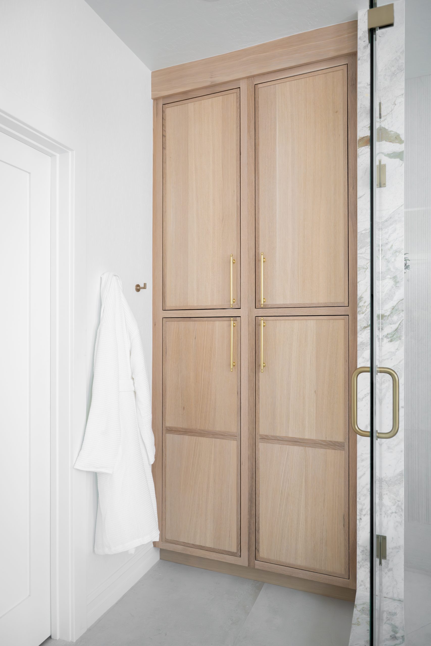Floor-to-ceiling white oak shaker linen tower with brass bar pulls and a robe hook beside the marble shower at The Charleston Southern Highlands remodel primary bathroom.