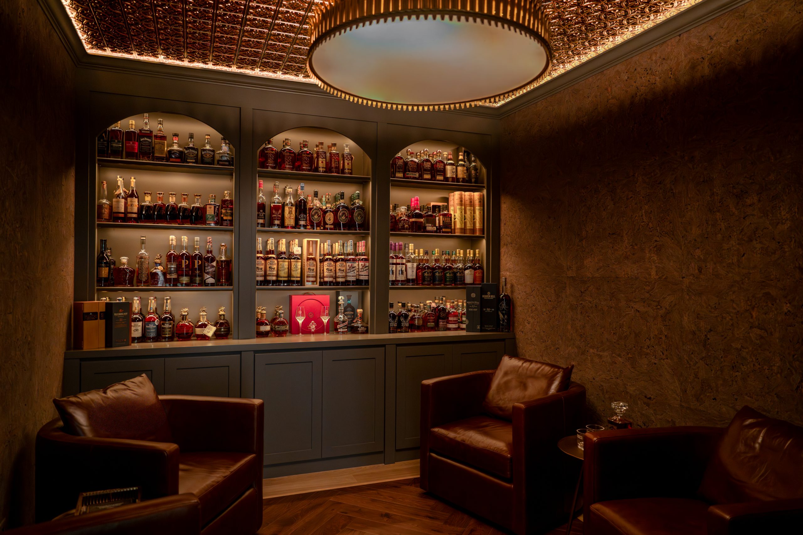 Custom whiskey lounge with three arched built-in liquor shelves, ornate pressed-metal ceiling, herringbone wood floor, and brown leather club chairs at The Charleston Southern Highlands remodel by Kingdom & Co.