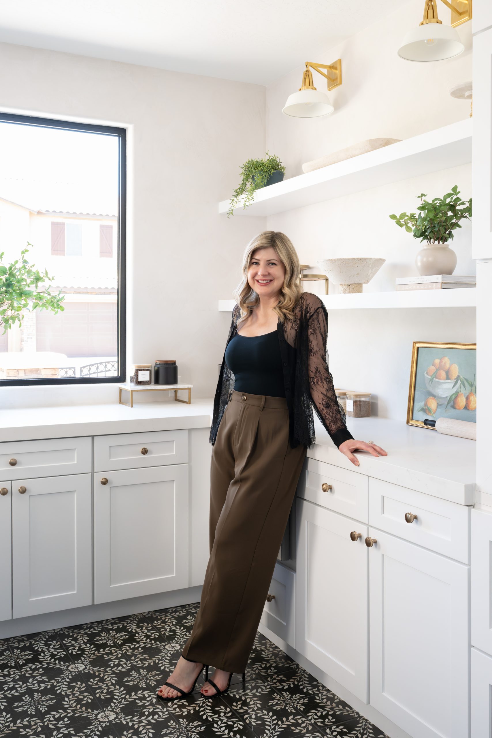 Jenn Coots, lead designer at Kingdom & Co. and the designer of The Charleston, in the project's scullery featuring white shaker cabinetry, brass wall sconces, open shelves with botanical art, and patterned floor tile at this Southern Highlands remodel.