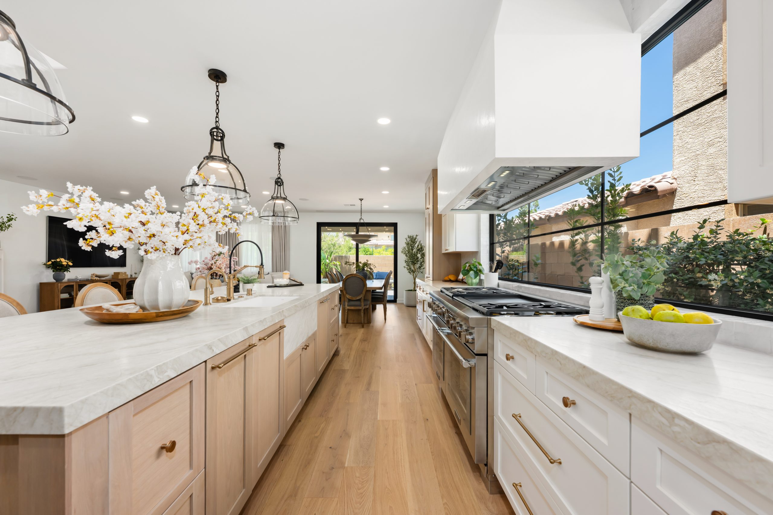 Wide view of The Charleston's kitchen featuring a long white oak shaker island, white marble countertops, glass dome pendants, custom plaster range hood, and steel-framed windows opening to the Southern Highlands courtyard remodel.
