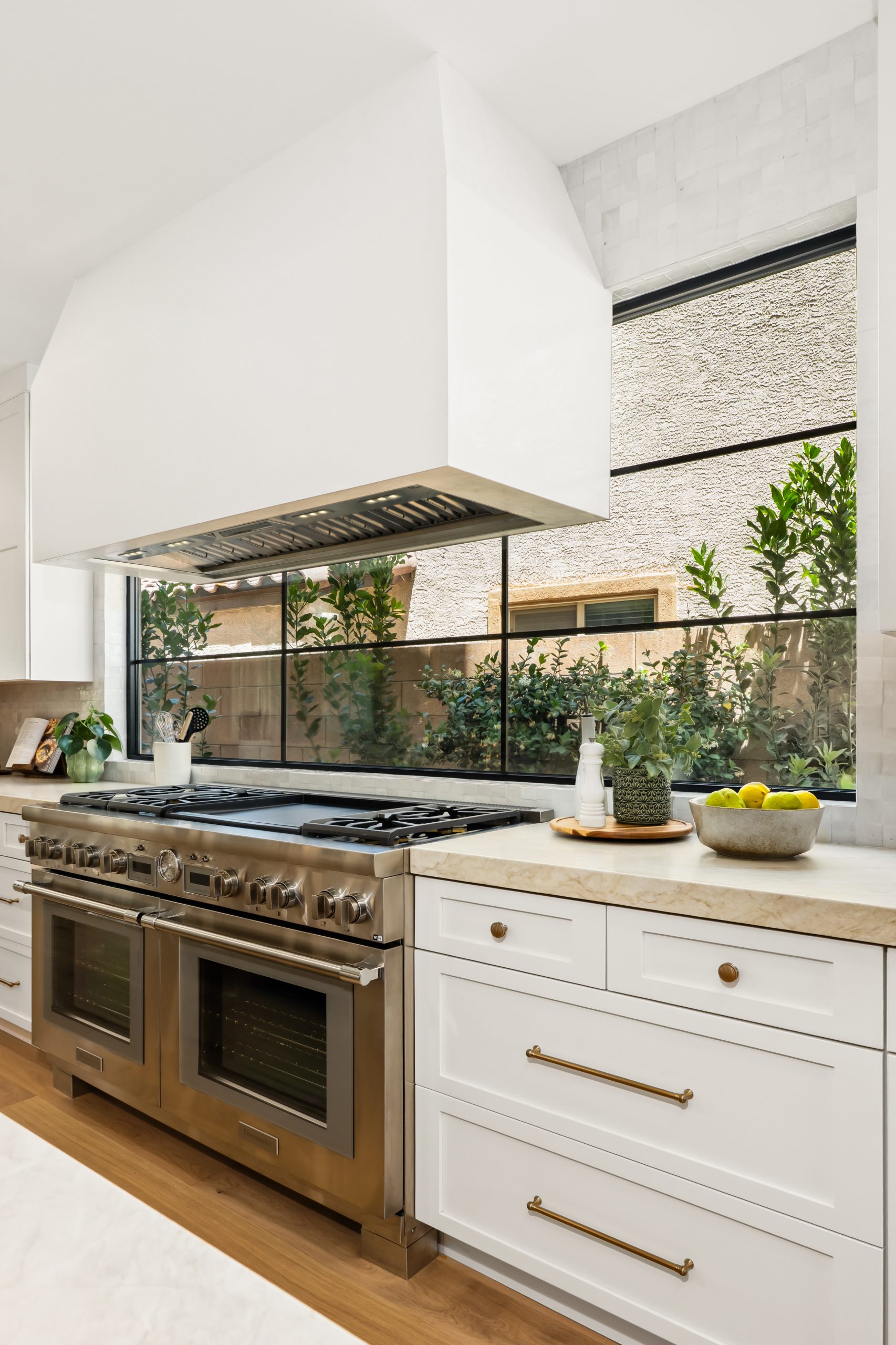 The Charleston_089 Professional dual-oven range with custom plaster hood, marble countertops, white shaker cabinetry, and steel-framed window opening to a desert courtyard at The Charleston Southern Highlands remodel.
