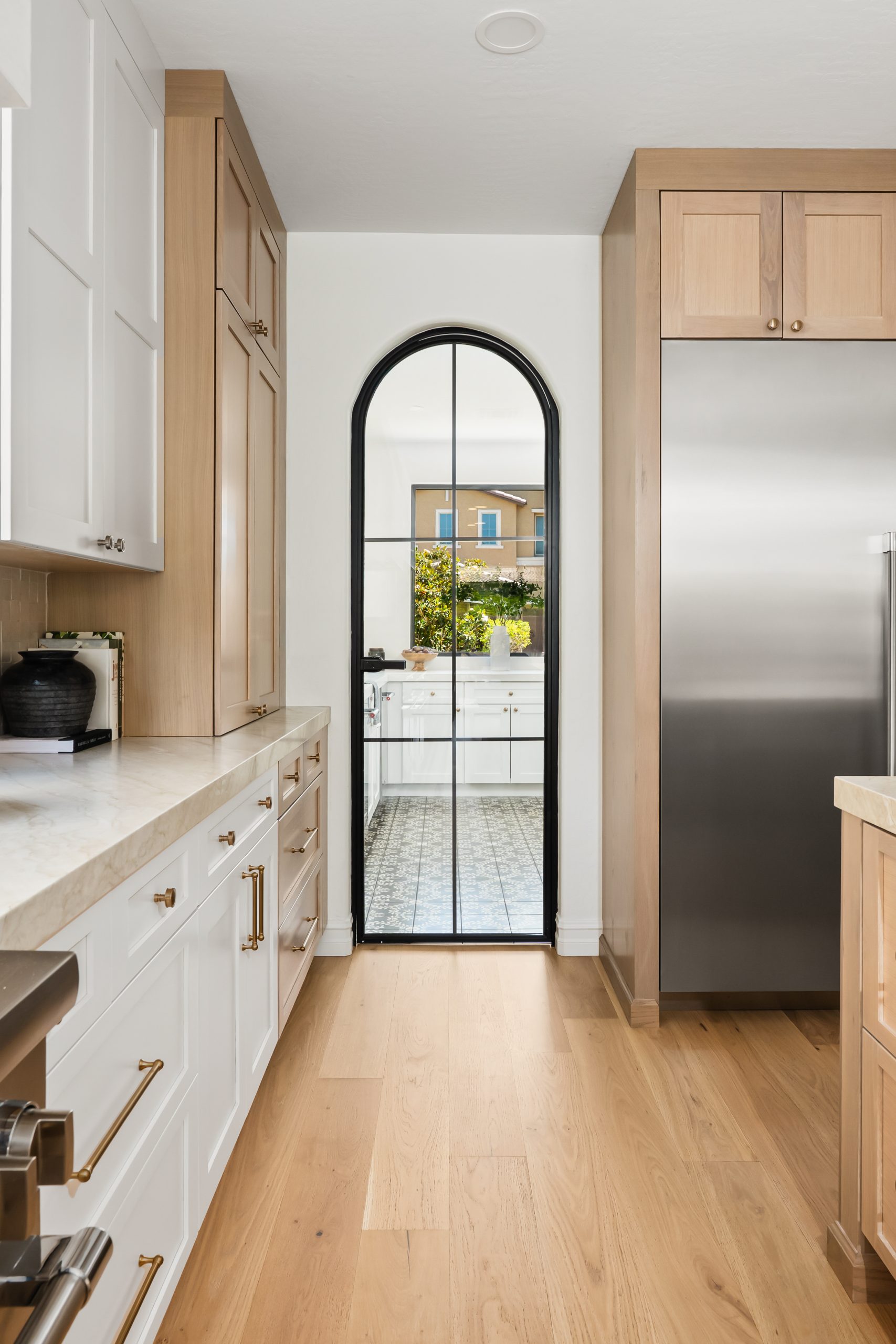 The Charleston_093 View into the butler's pantry through an arched black steel and glass door, framed by white oak shaker cabinetry, white marble countertops, and brass hardware at The Charleston Southern Highlands remodel.