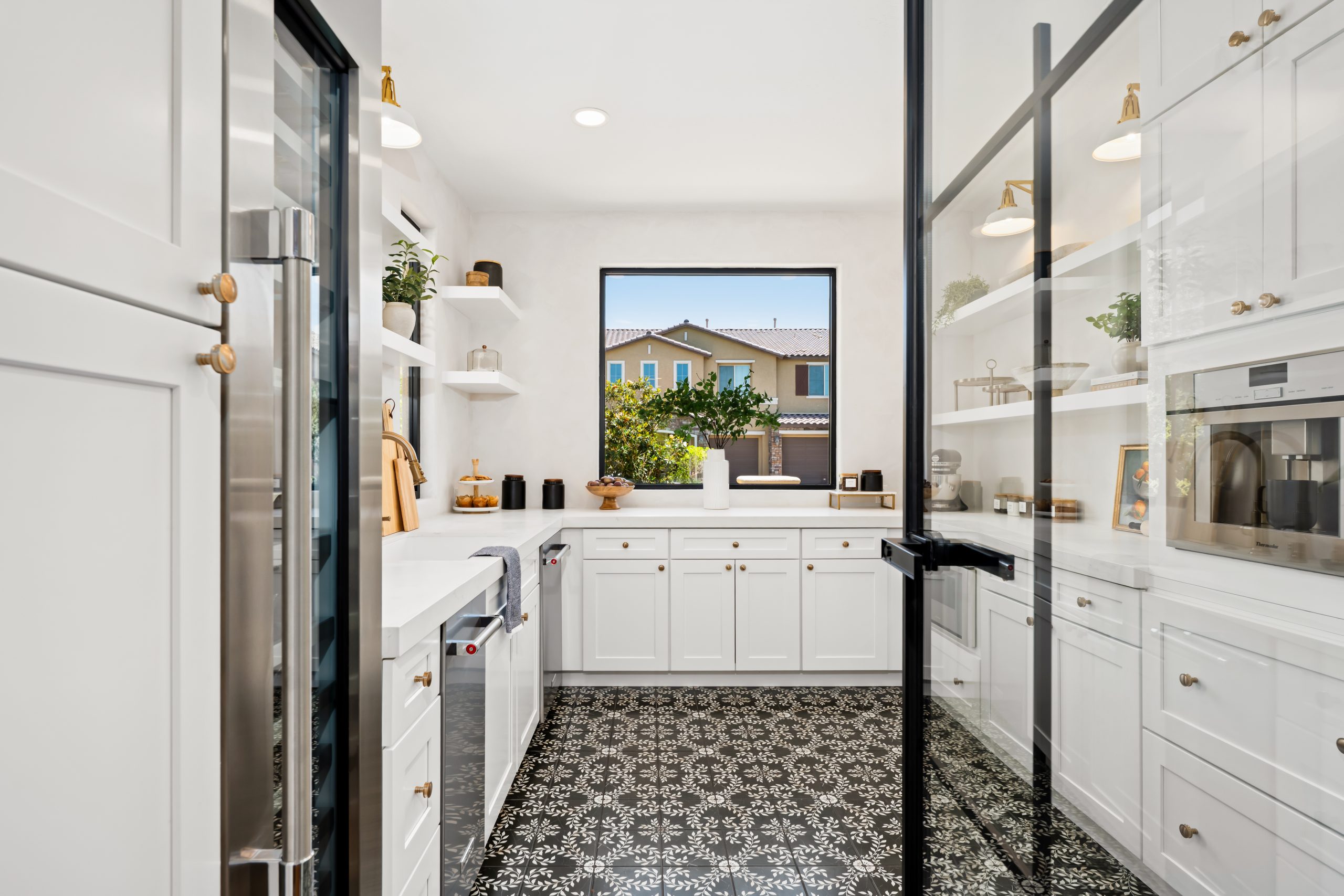View into the walk-in scullery through black steel and glass doors, featuring white shaker cabinetry, intricate black and white patterned floor tile, built-in coffee station, and brass wall sconces at The Charleston Southern Highlands remodel.