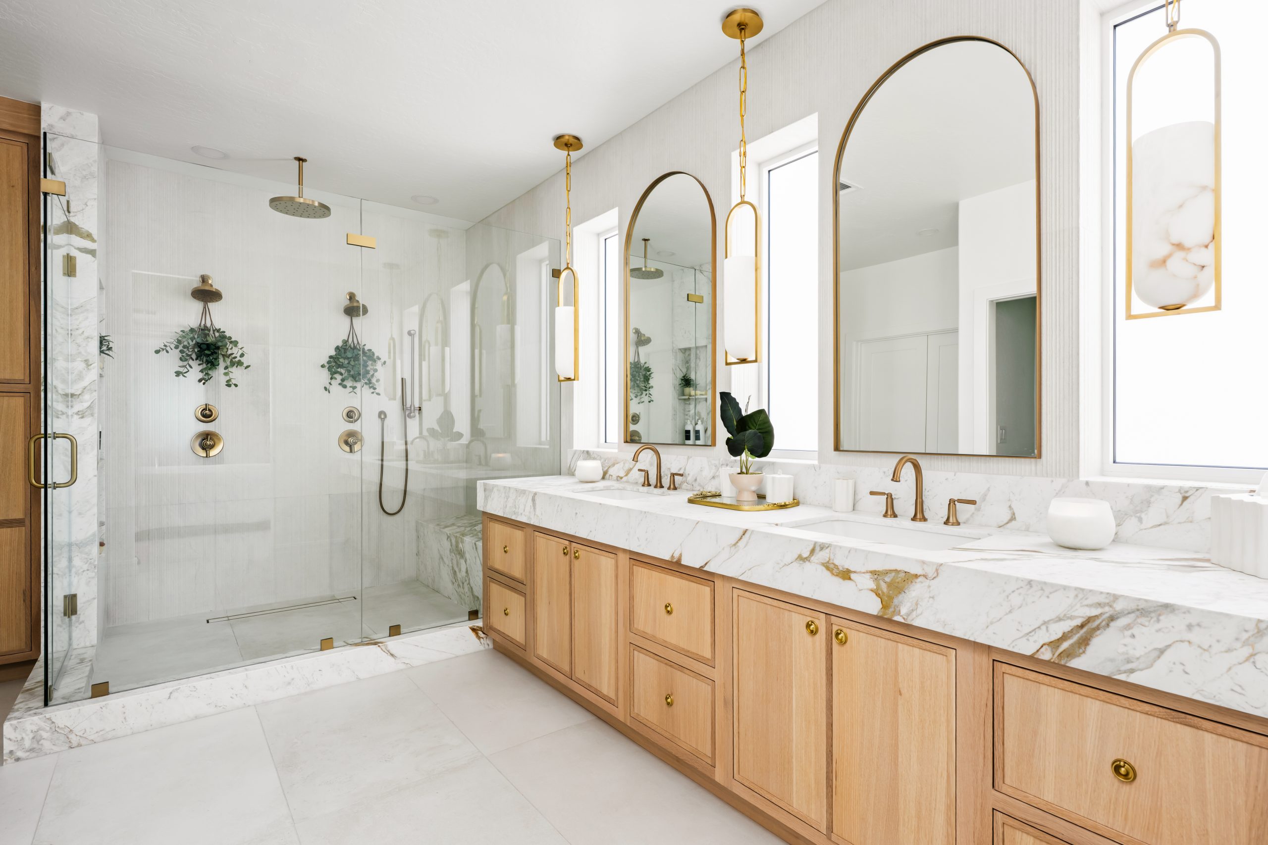 Primary bathroom with glass-enclosed walk-in shower, calacatta marble surround, double vanity with arched brass mirrors, alabaster pendants, and white oak shaker cabinetry at The Charleston Southern Highlands remodel.