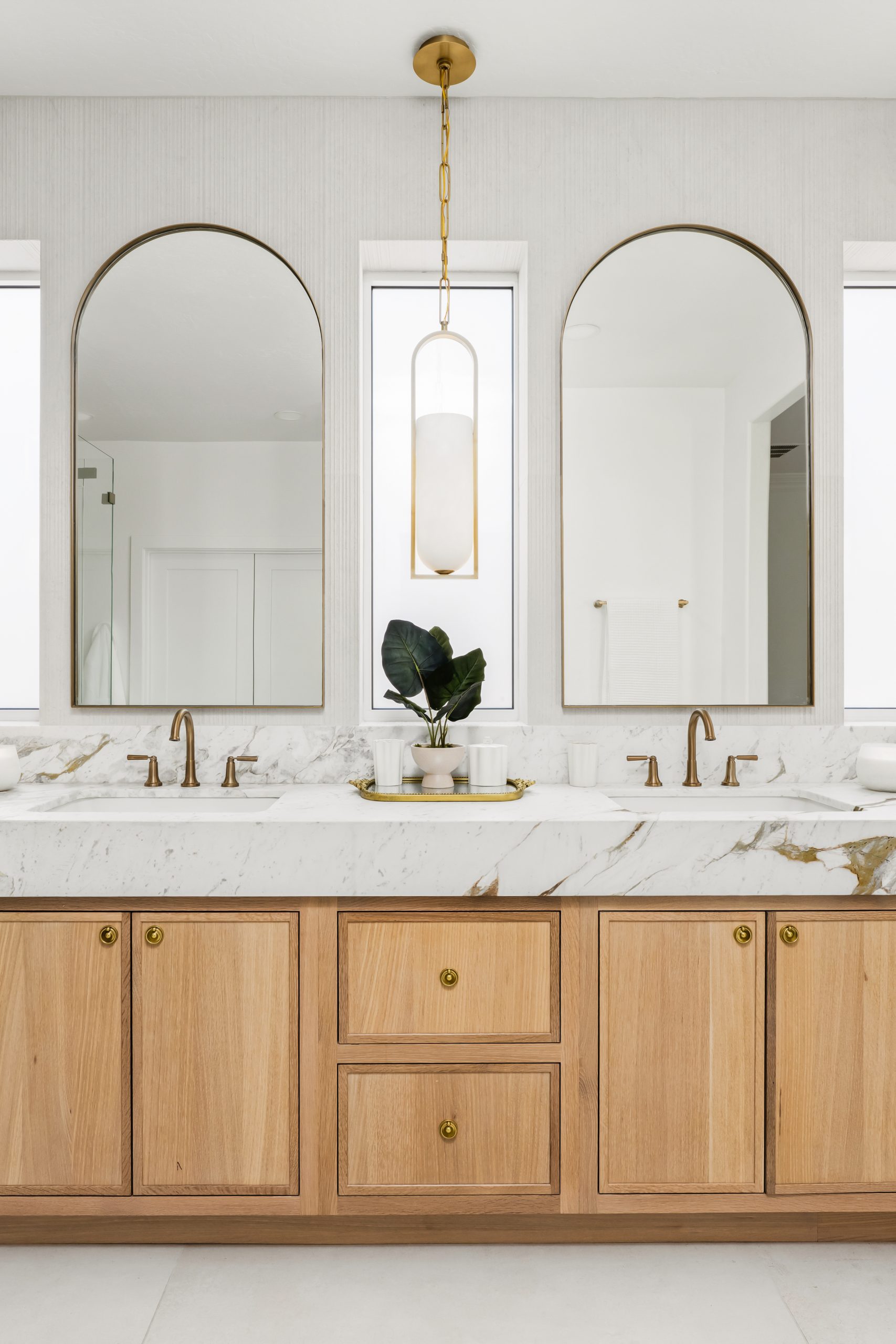 Primary bathroom double vanity with twin brass-framed arched mirrors, white oak shaker cabinetry, calacatta marble waterfall countertop, and a central brass pendant at The Charleston Southern Highlands remodel.