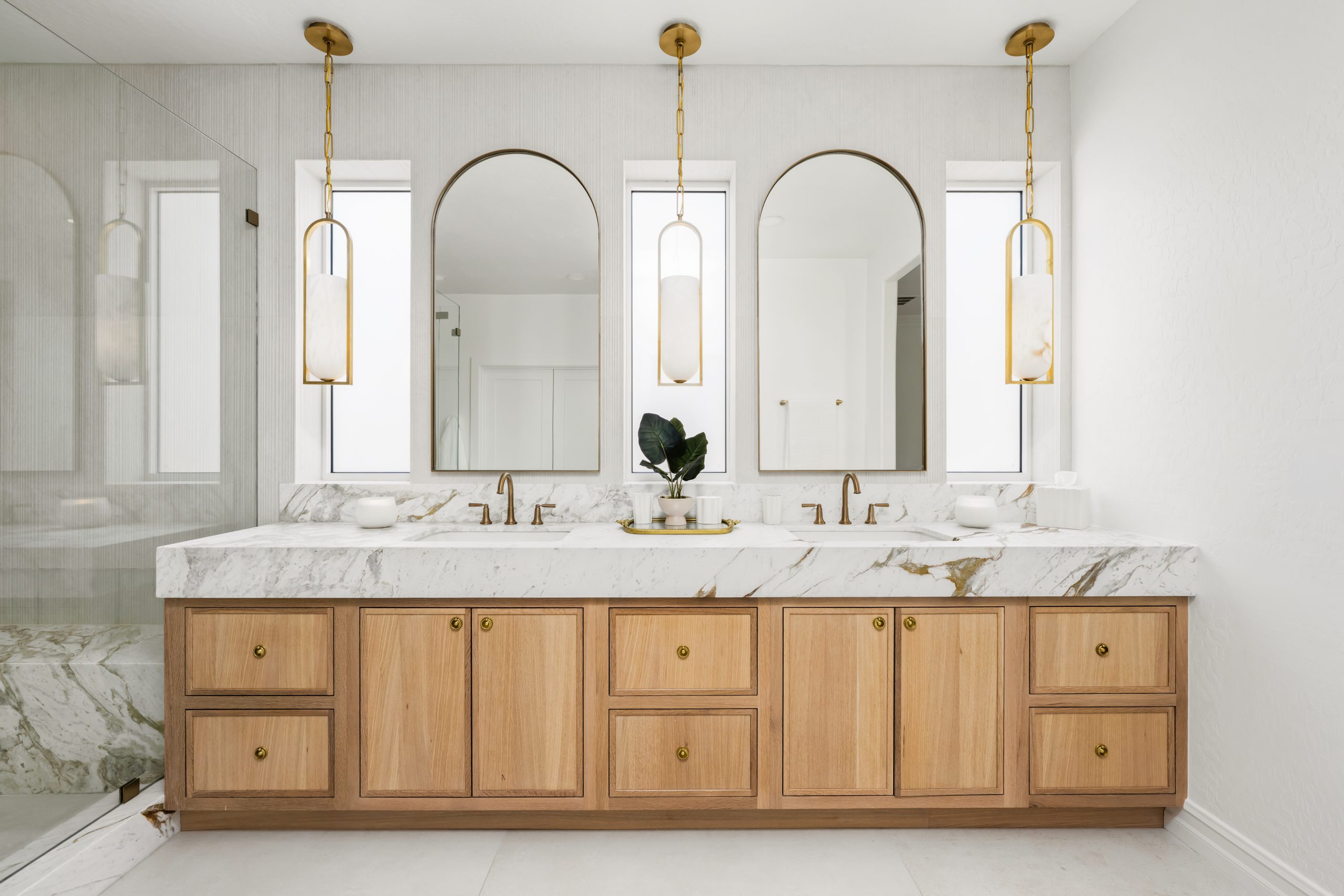 Primary bathroom double vanity with two arched brass-framed mirrors, three alabaster pendants, white oak shaker cabinetry, and calacatta marble waterfall countertop at The Charleston Southern Highlands remodel.