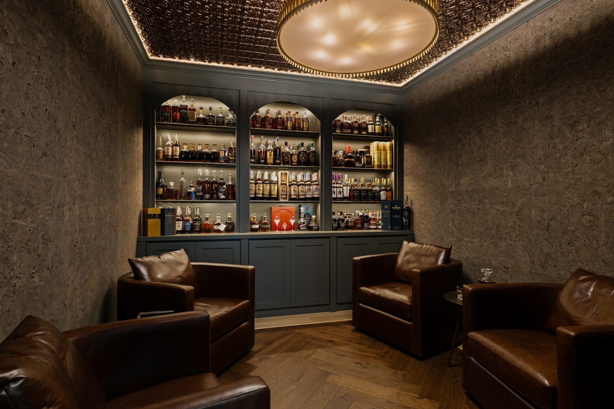 Wide view of the custom whiskey lounge with three arched bourbon displays, four brown leather club chairs, pressed metal ceiling with central round cove, cork-textured walls, and herringbone wood floor at The Charleston Southern Highlands remodel.