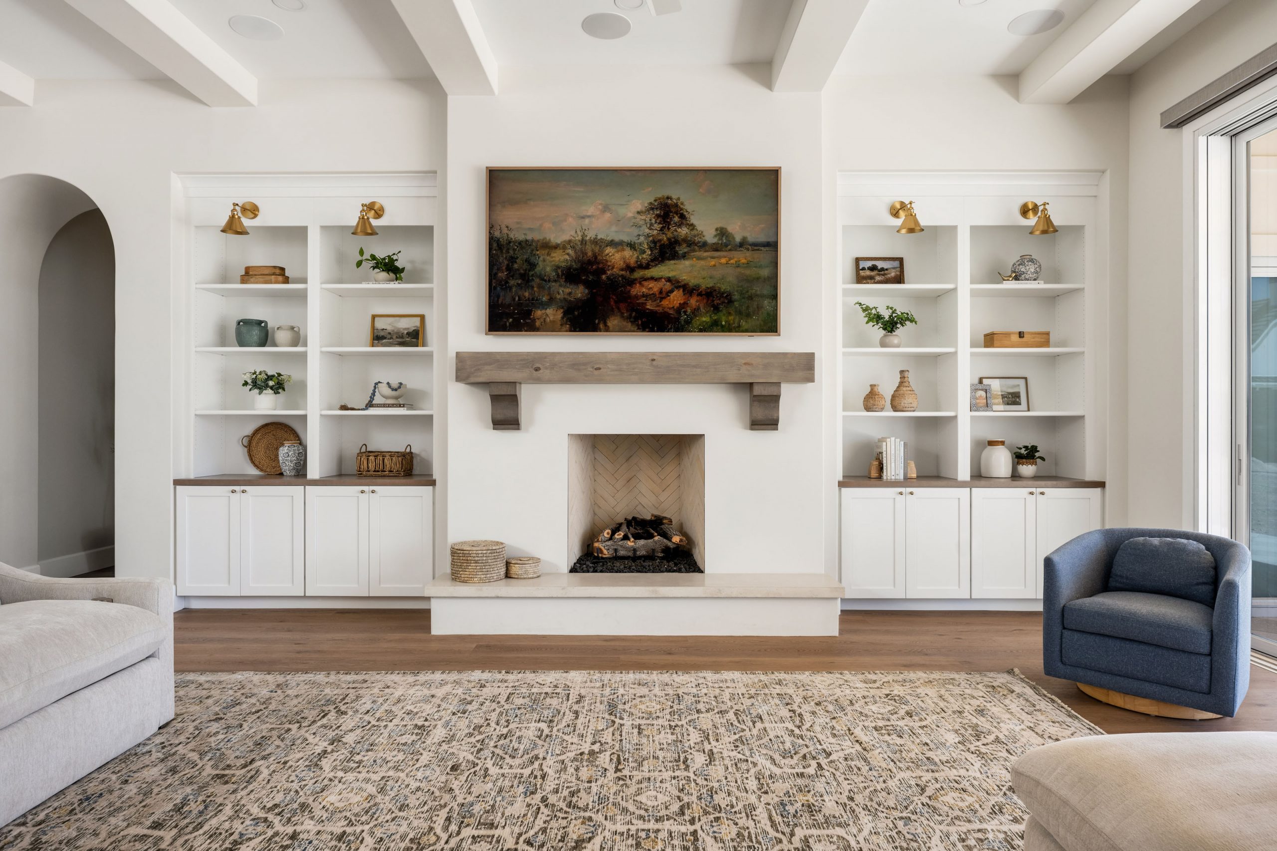 White Gables Estate002 Great room with white built-in shelving flanking a stone fireplace, reclaimed wood beam mantel, herringbone firebox, landscape painting, brass sconces, and faux ceiling beams — The White Gables Estate cottage style custom home in Centennial Hills, Las Vegas by Kingdom & Co.