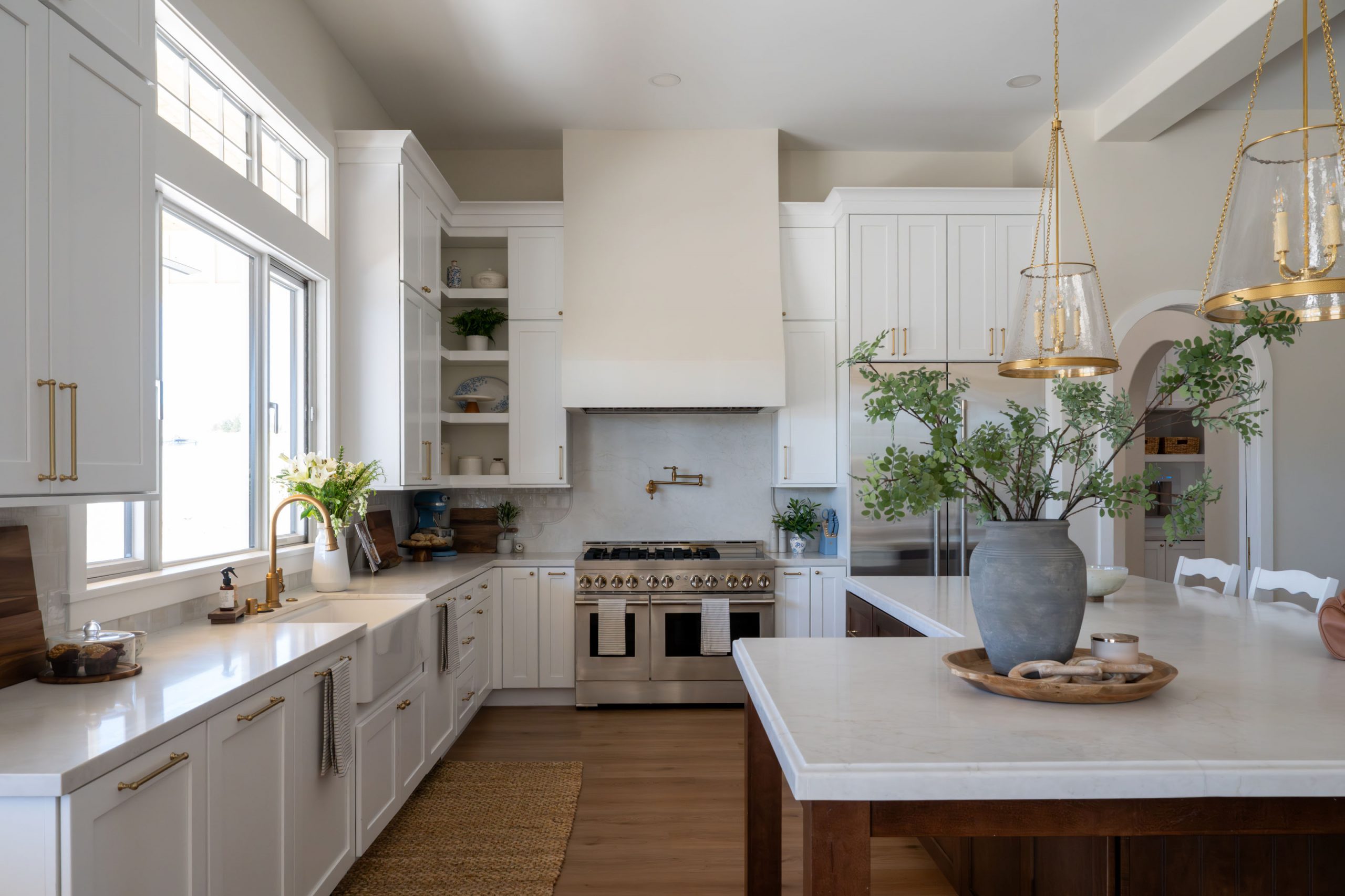 Cook's kitchen view with white range hood, stainless dual-oven range, brass pot filler, farmhouse apron sink, brass faucet, brass pendant lanterns, and walnut-stained island — The White Gables Estate cottage style custom home Las Vegas by Kingdom & Co