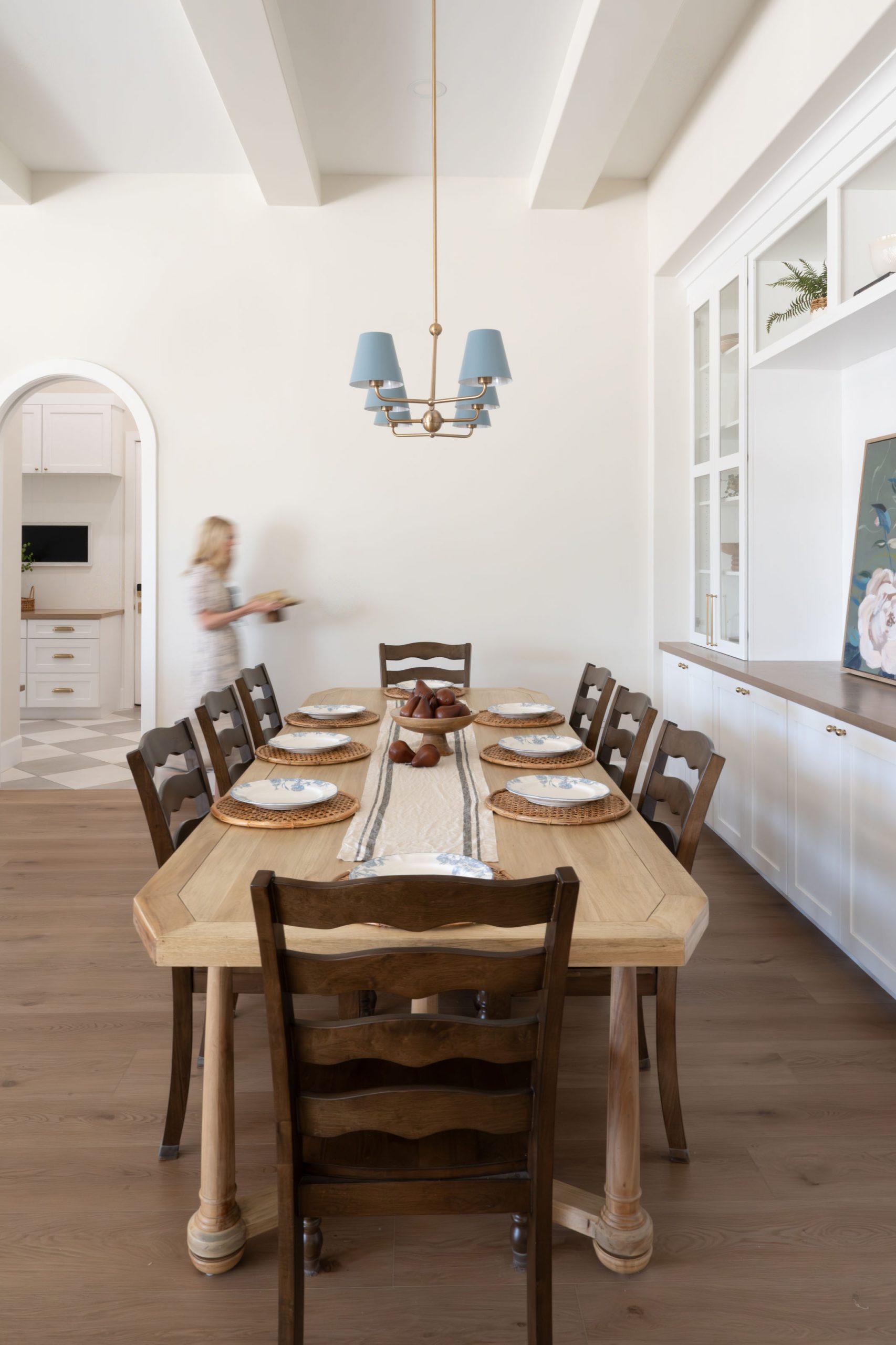 Dining room with white faux ceiling beams, blue-shaded chandelier, ladderback chairs, and archway leading to butler's pantry with checkerboard tile floor — The White Gables Estate cottage style custom home in Centennial Hills, Las Vegas by Kingdom & Co.
