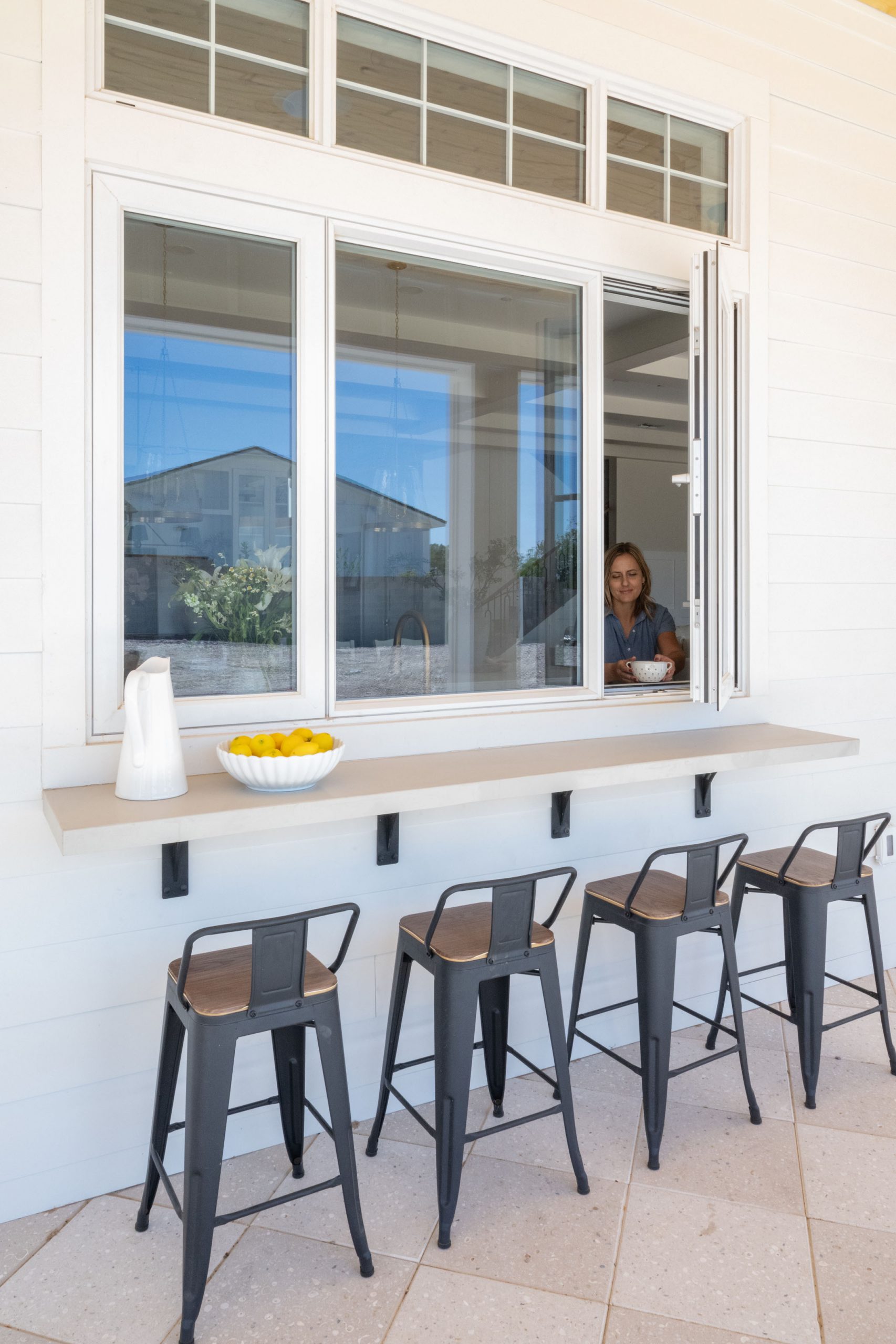Outdoor patio with kitchen pass-through window counter, black metal counter stools, white painted exterior, and transom windows — The White Gables Estate by Kingdom & Co.