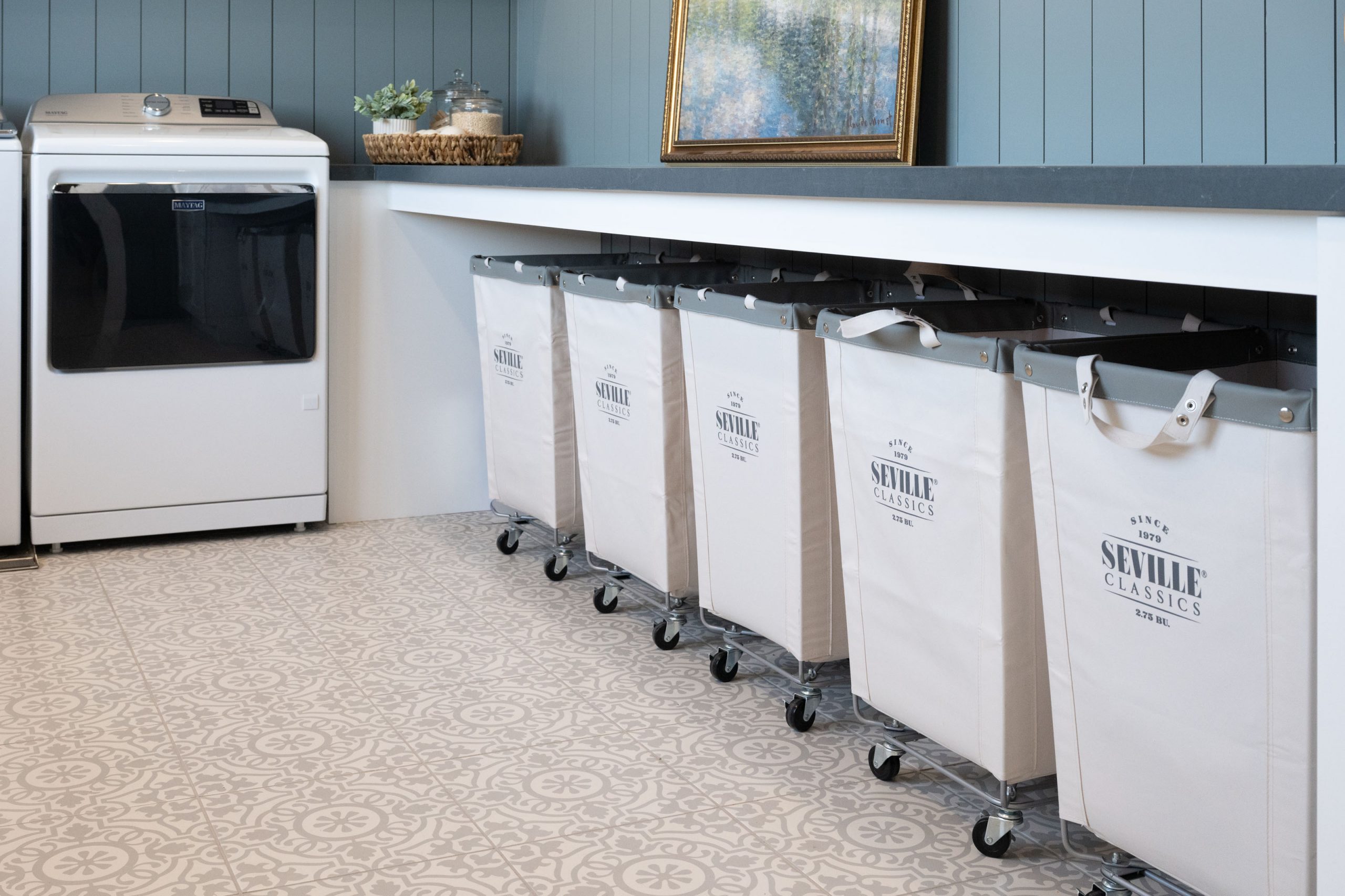 Custom laundry room with patterned cement-look tile floor, painted shiplap walls, framed landscape art, washer/dryer, and five rolling commercial laundry sorters built for a family of seven — The White Gables Estate cottage style custom home Las Vegas by Kingdom & Co.