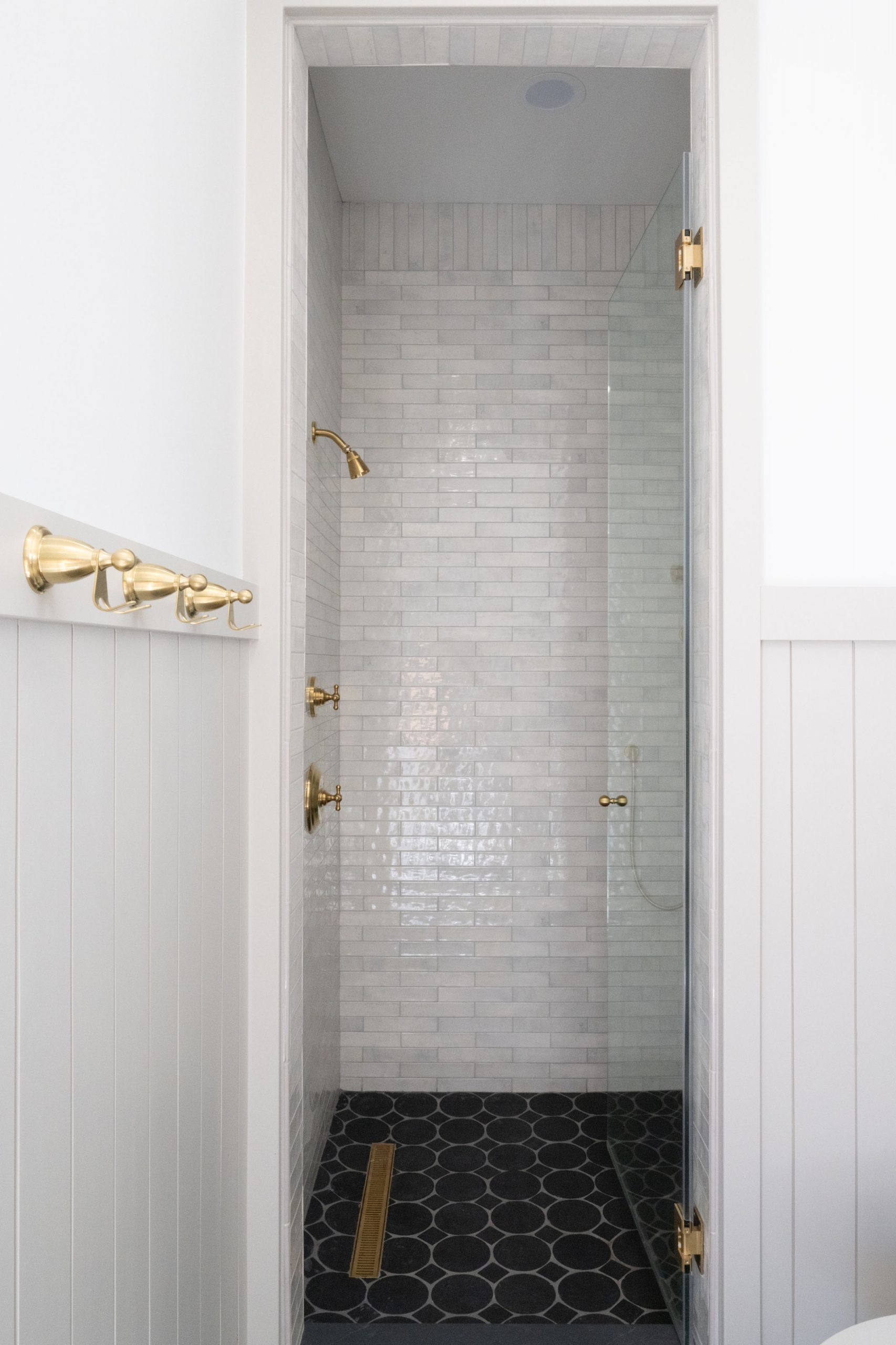 Guest bathroom shower with handmade-look subway tile walls, brass shower head, brass body sprays, black hex mosaic floor, brass linear drain, and white shiplap wainscoting — The White Gables Estate by Kingdom & Co.