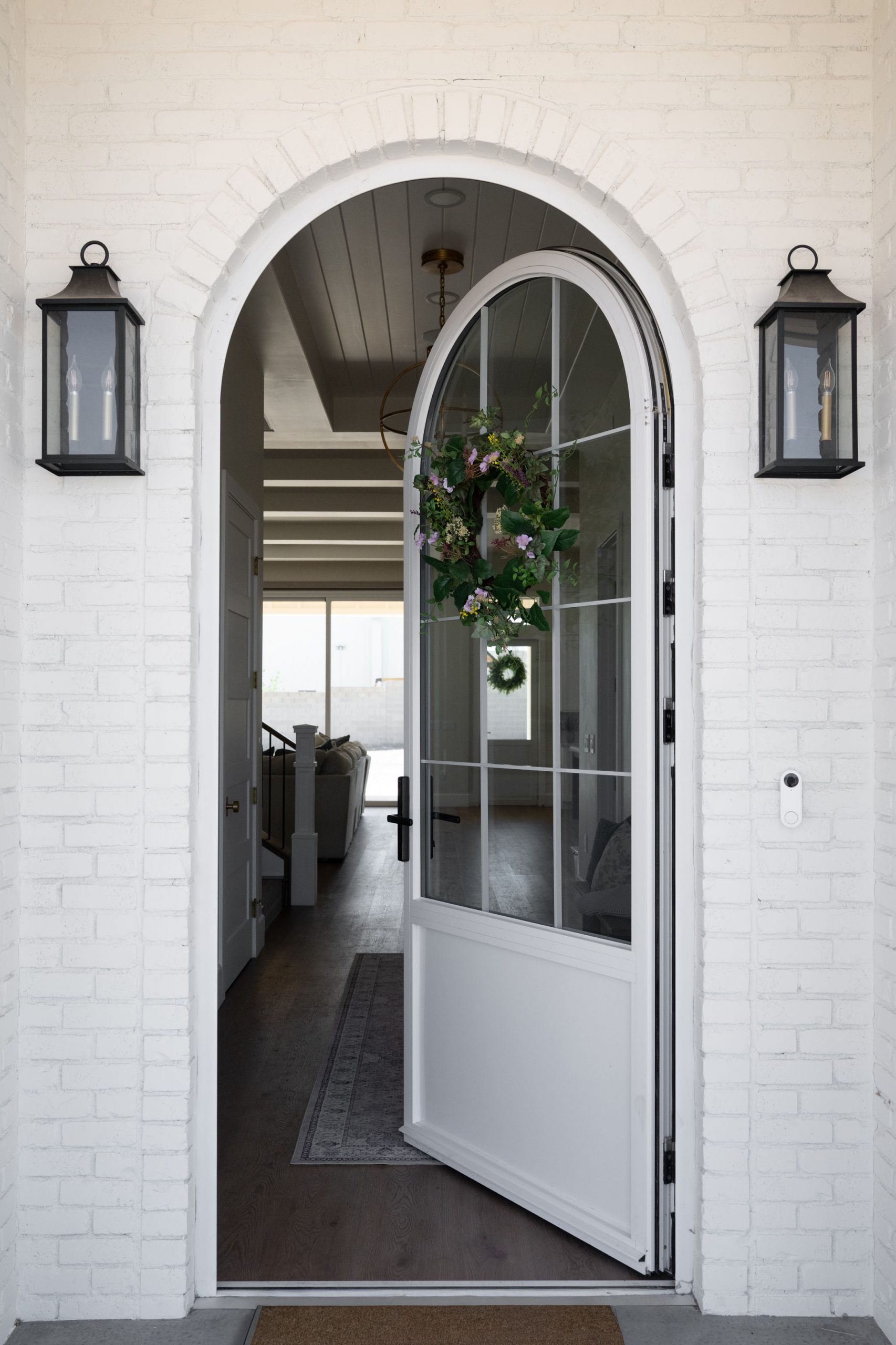 White Gables Estate117 Arched front entry with white painted brick exterior, black coach-style lantern sconces, and arched glass-and-panel front door with floral wreath — The White Gables Estate cottage style custom home in Centennial Hills, Las Vegas by Kingdom & Co.