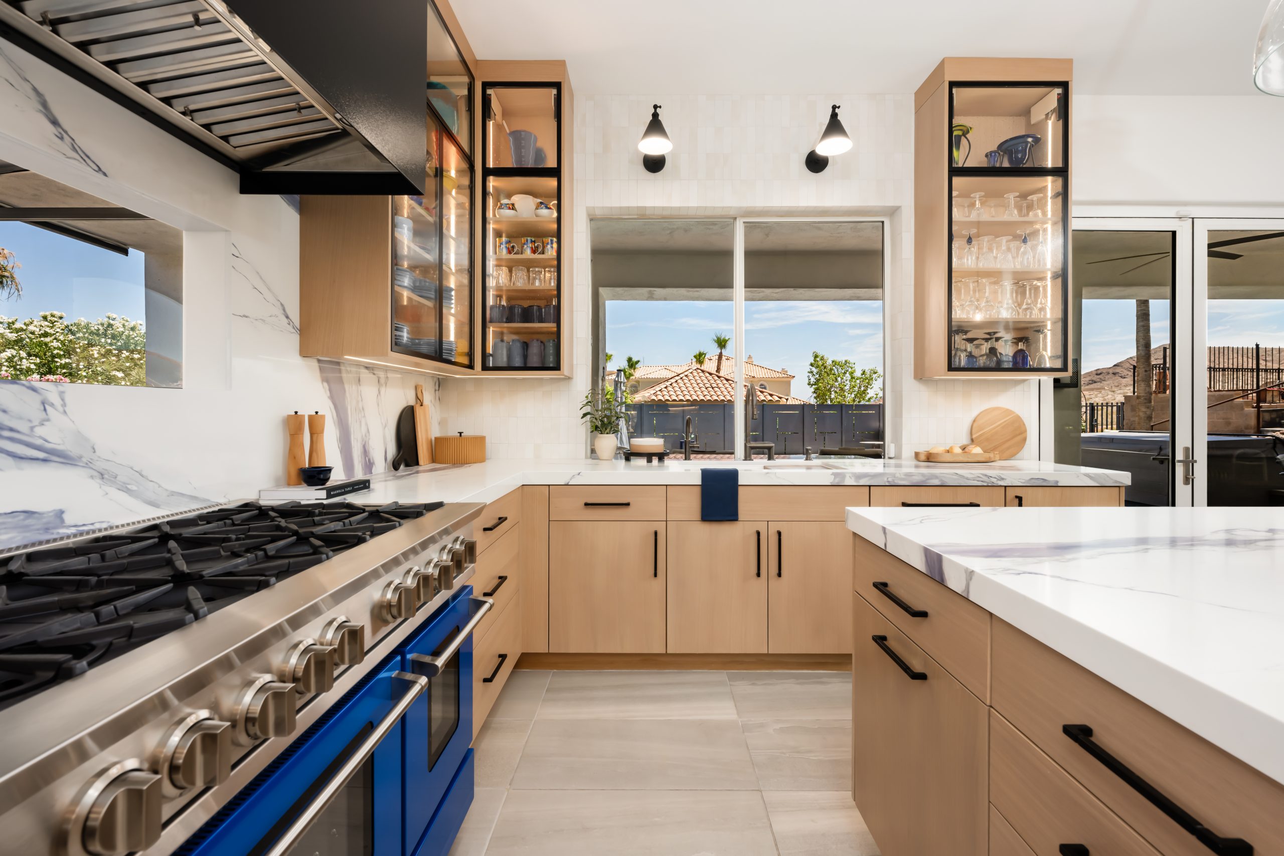 Kitchen with a cobalt blue dual-oven range, blue-veined marble slab backsplash, black range hood, and natural white oak cabinetry with black hardware at The Azul Ridge in Boulder City.