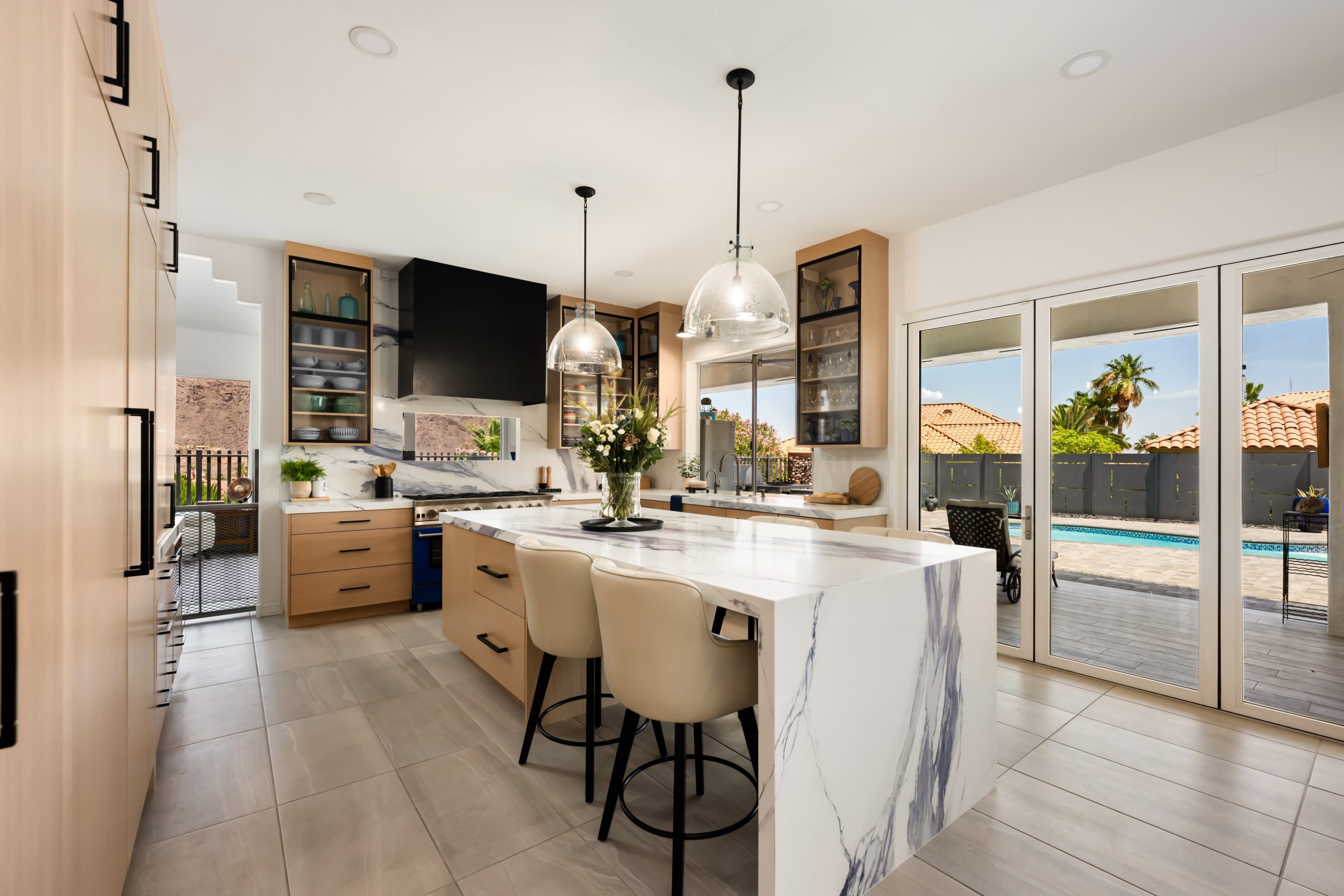 Kitchen with a blue-veined marble waterfall island, cream leather barstools, glass dome pendants, and floor-to-ceiling sliding doors opening to the pool at The Azul Ridge in Boulder City.