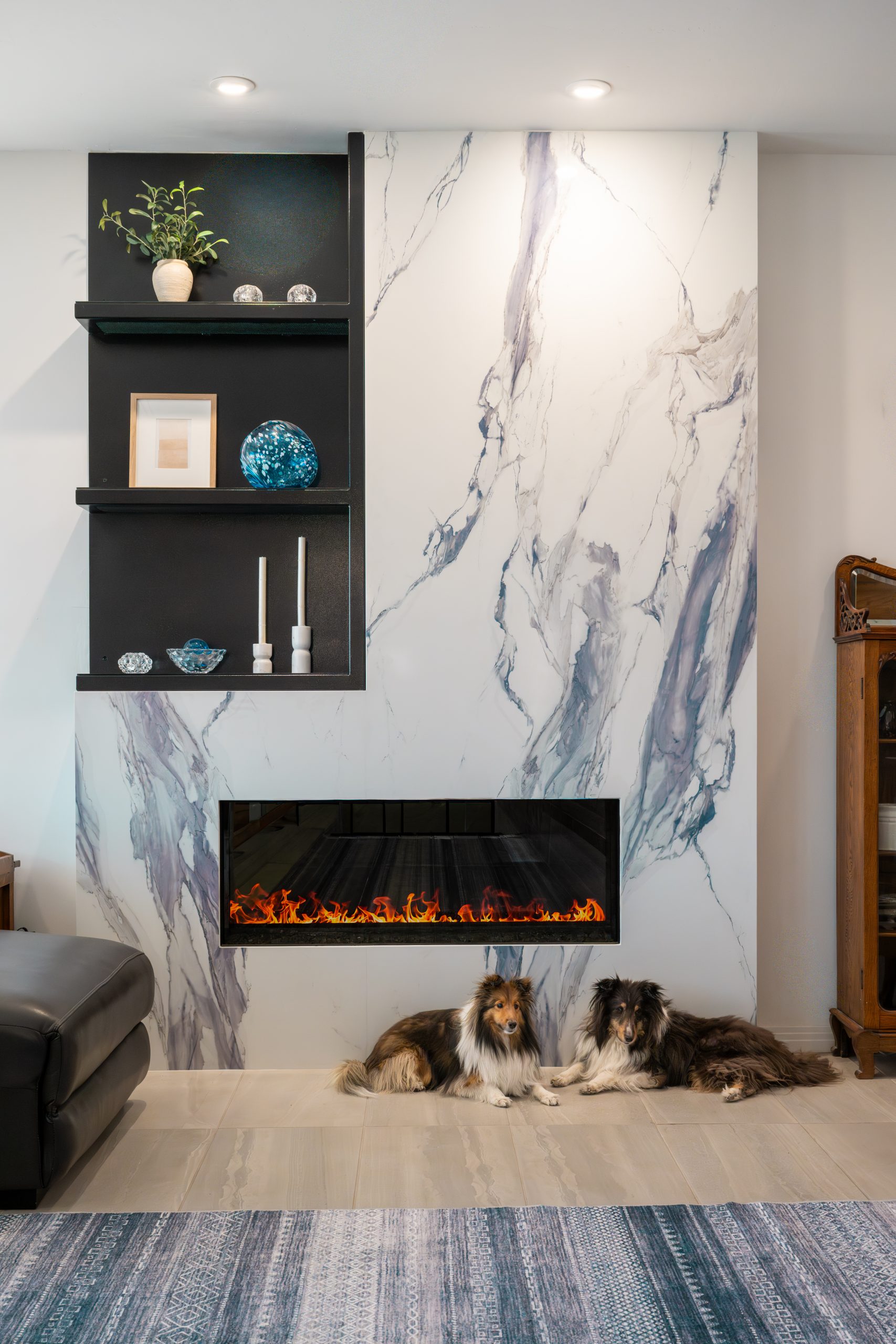 Living room fireplace wall with a linear electric fireplace, blue-veined marble slab surround, built-in black shelving, and two Shetland sheepdogs on the tile floor at The Azul Ridge in Boulder City.