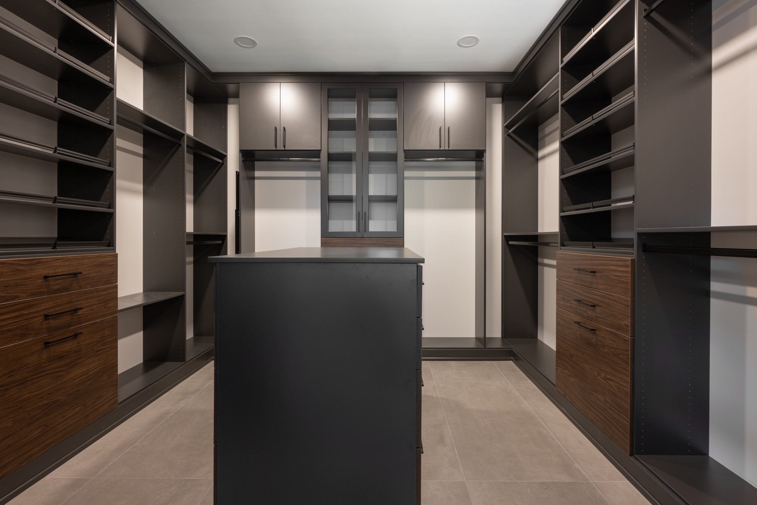 Primary walk-in closet with dark charcoal built-ins, walnut drawers, and center dresser island, Kingdom & Co. The Lakes.
