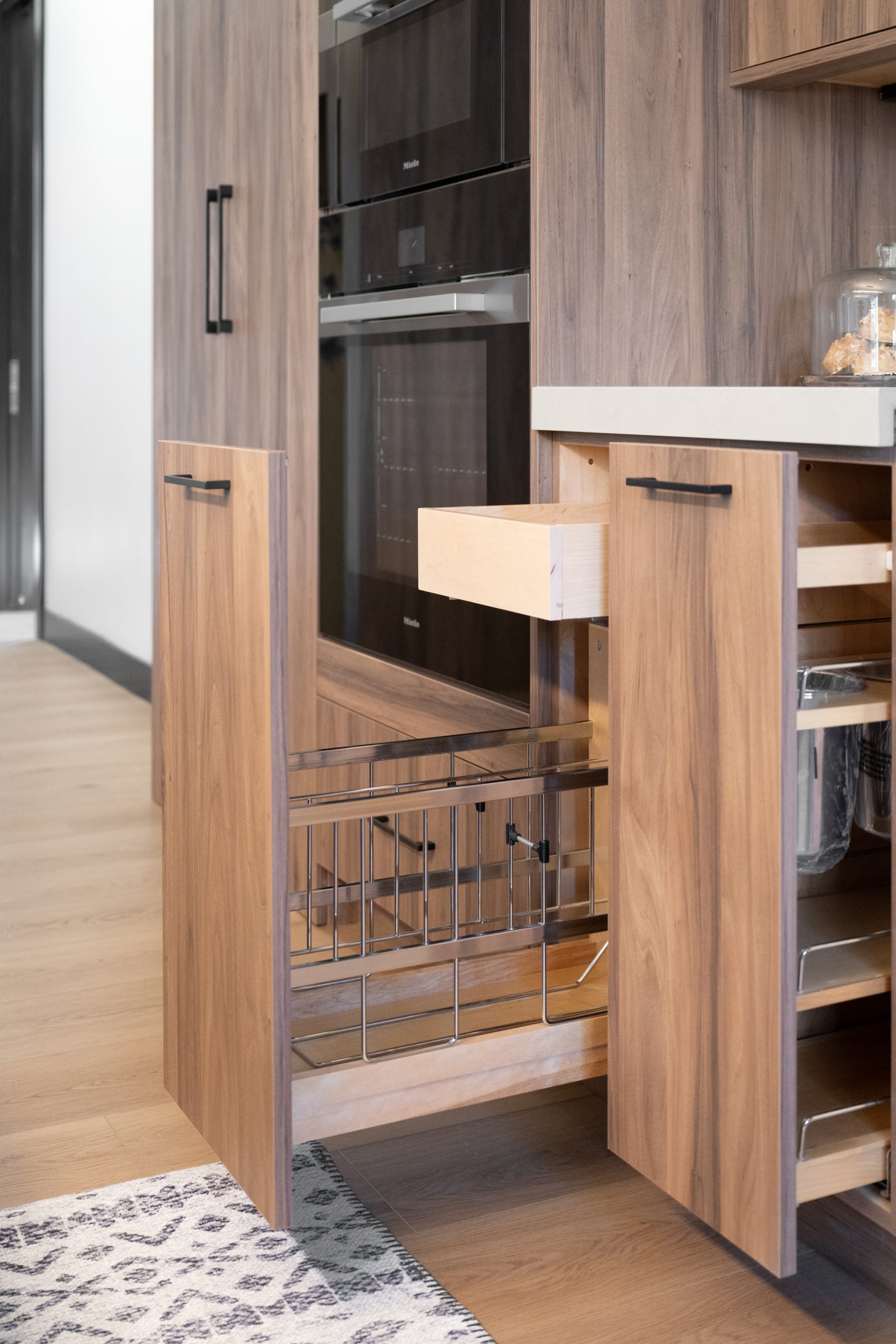 Pull-out pantry cabinet with wire baskets beside Miele wall ovens, Kingdom & Co. The Lakes kitchen.