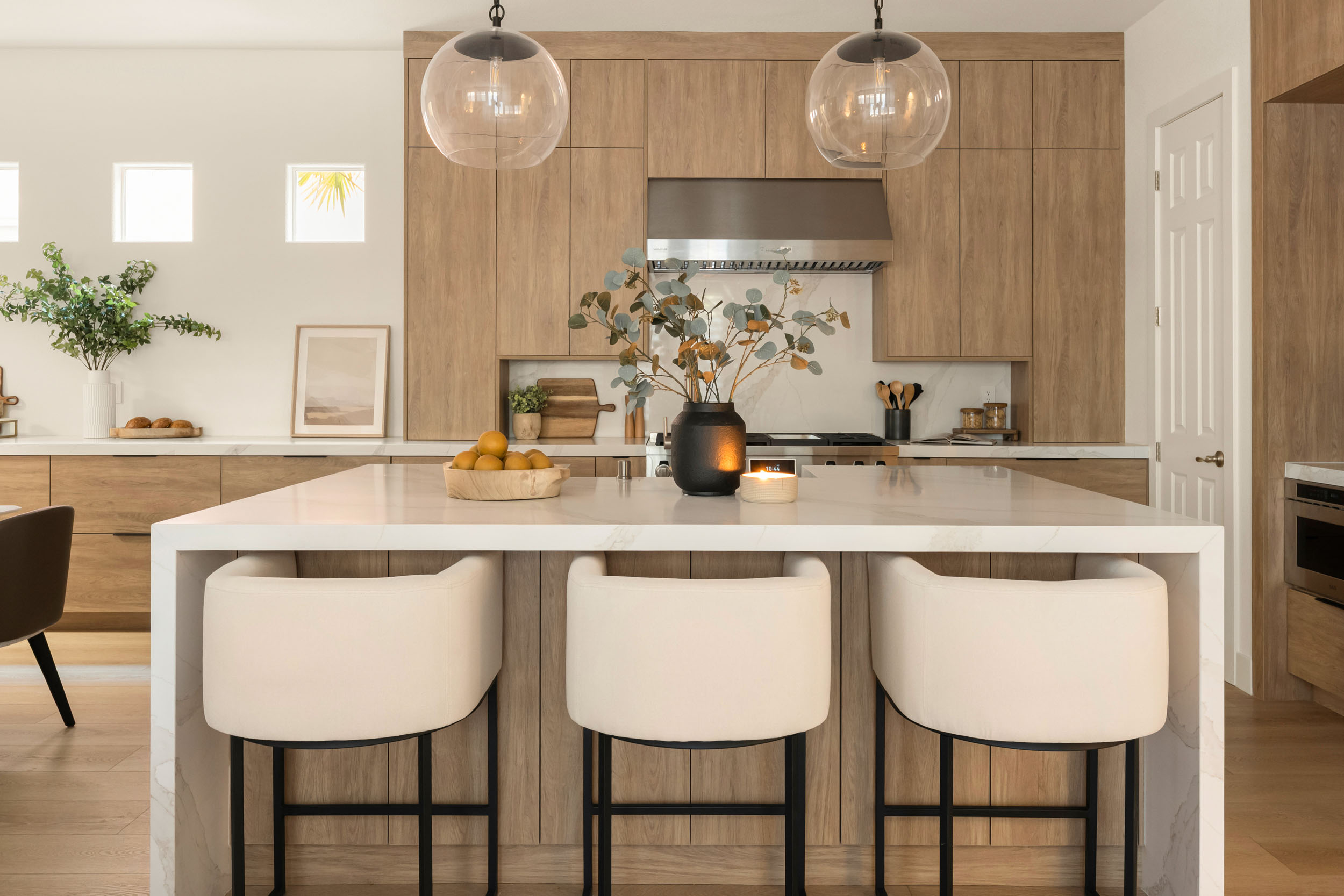 Warm oak kitchen elevation from dining with marble waterfall island, cream leather counter stools, and glass globe pendants in Summerlin remodel by Kingdom & Co.