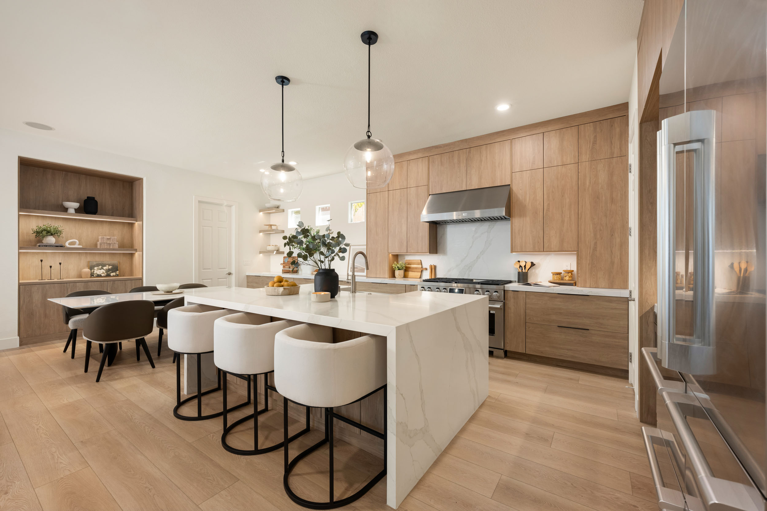 Warm oak kitchen wide-angle with marble waterfall island, cream leather counter stools on black steel legs, glass globe pendants, and floor-to-ceiling oak cabinetry framing the integrated stainless hood in Traccia Summerlin remodel by Kingdom & Co.