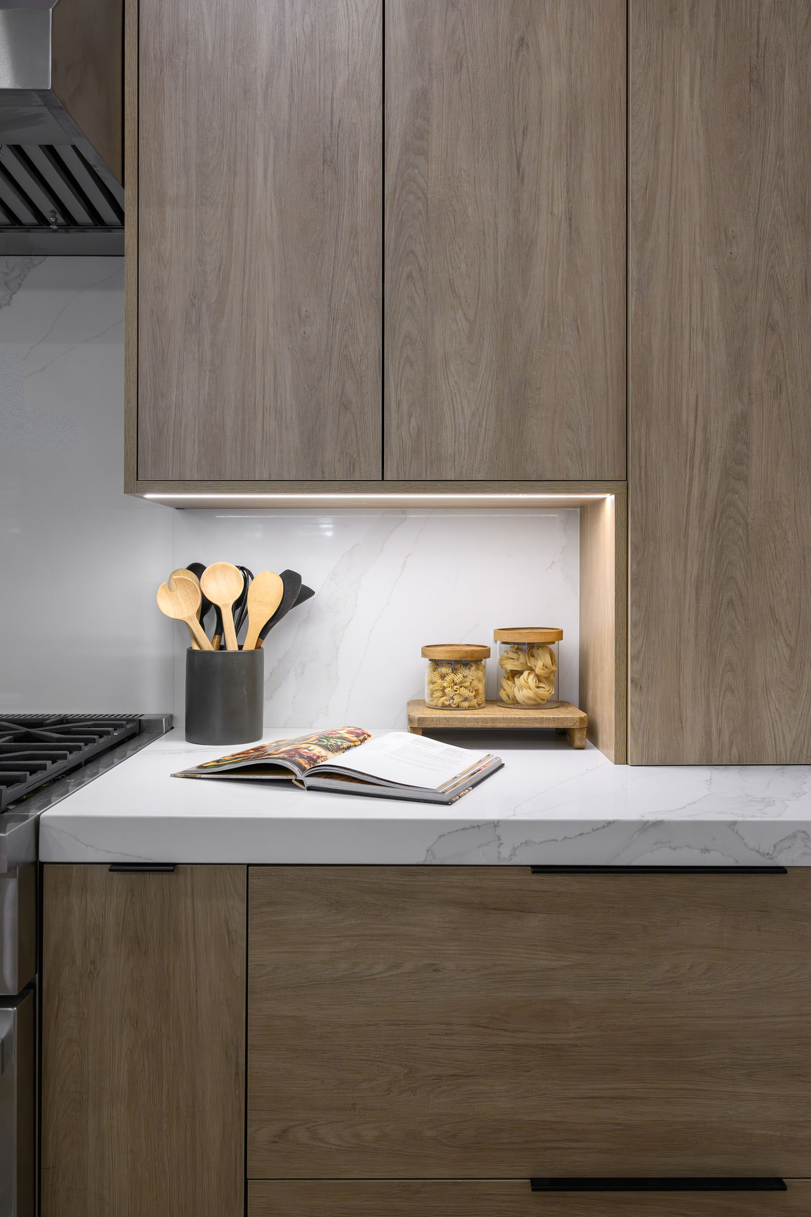 Warm oak flat-panel cabinets with slim black tab pulls and lit under-cabinet niche in Summerlin remodel kitchen by Kingdom & Co.