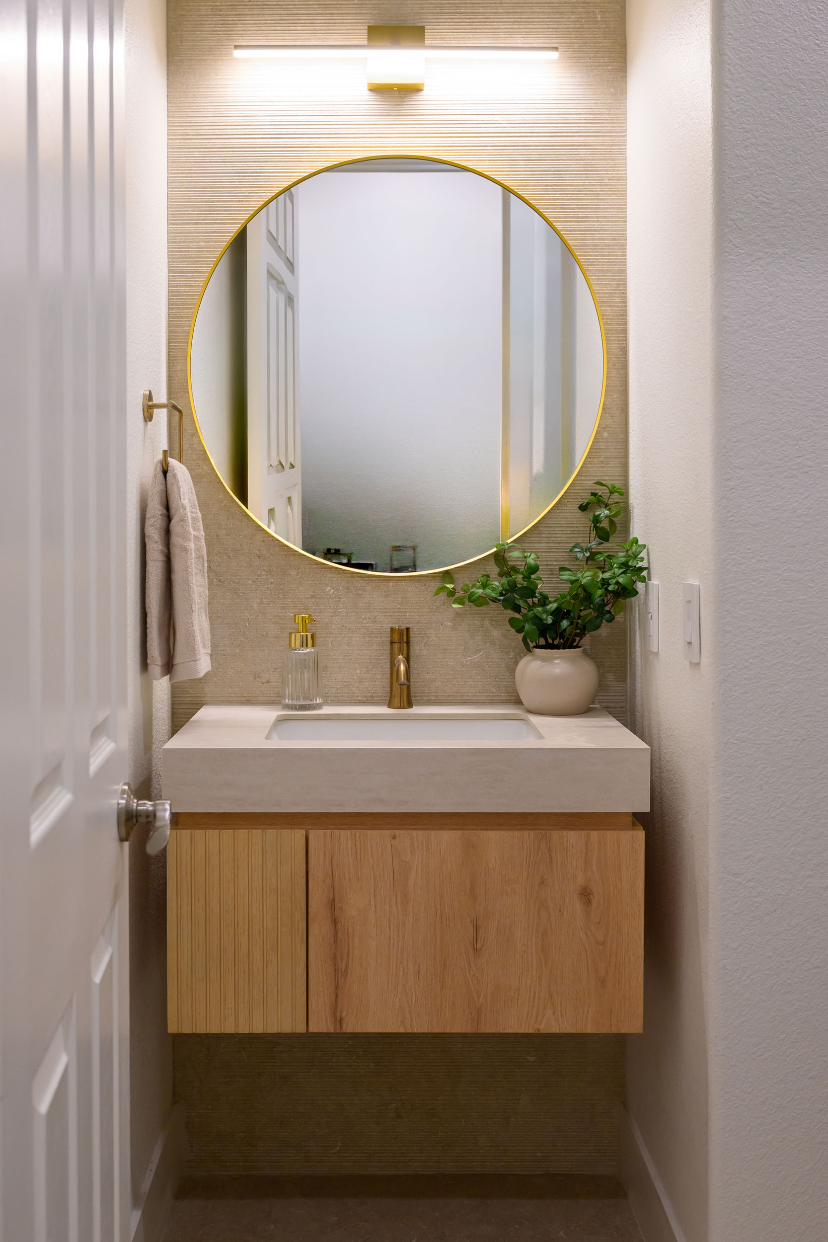 Powder room with fluted oak floating vanity, integrated stone sink, brass round mirror, and linear sconce in Summerlin remodel by Kingdom & Co.