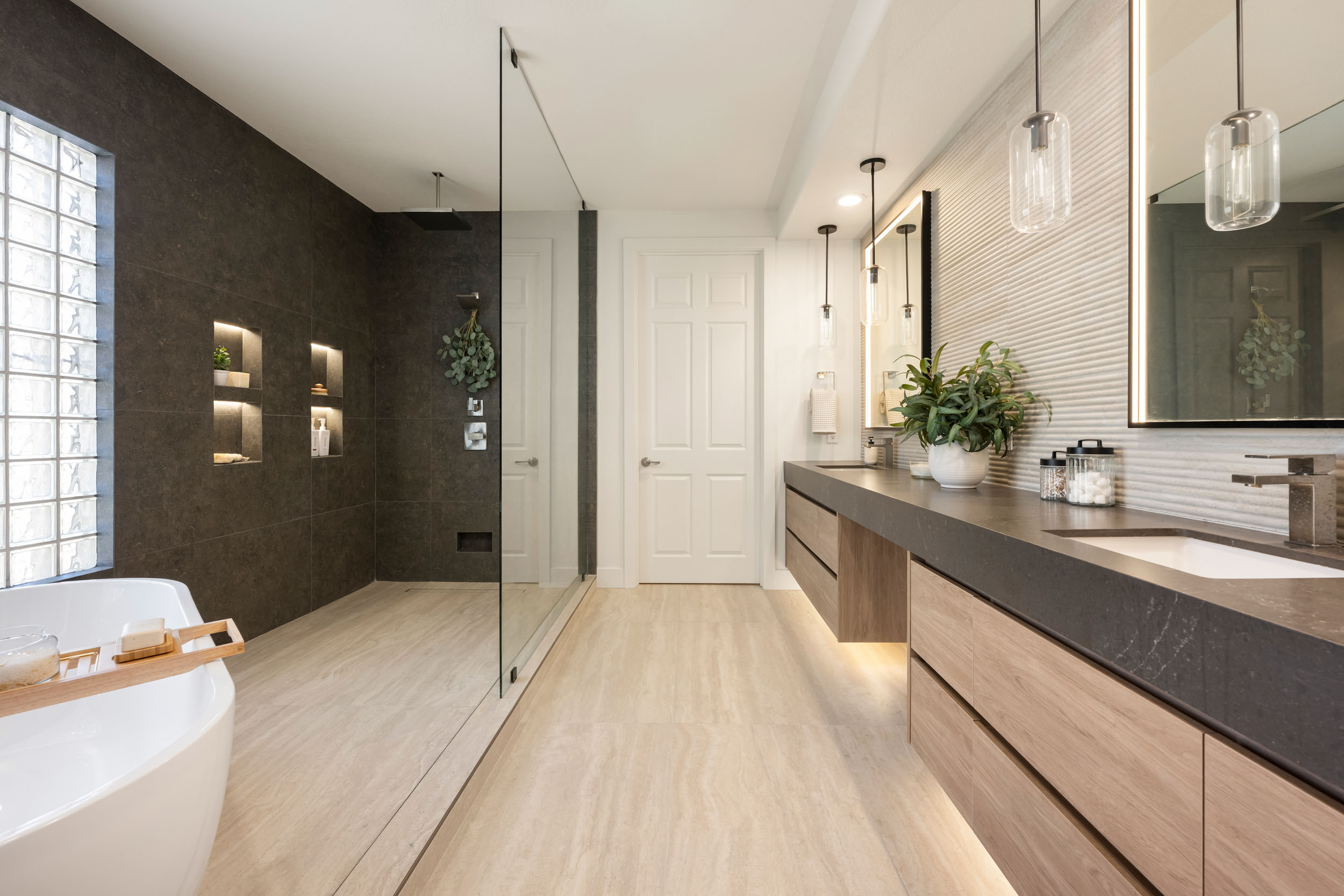 Primary bathroom full wide with freestanding soaker tub, curbless walk-in shower with dark charcoal stone accent wall, and long floating oak double vanity in Traccia Summerlin remodel by Kingdom & Co.