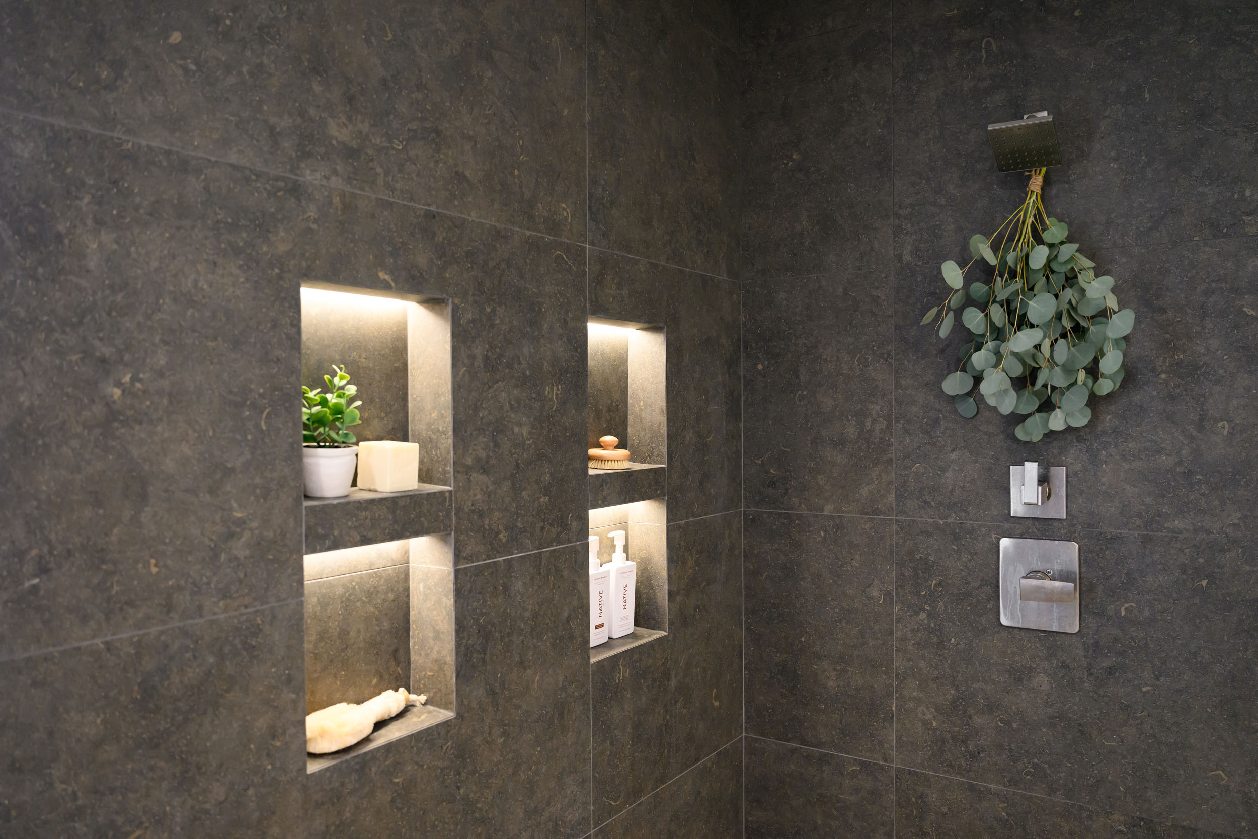 Primary shower detail with dark charcoal stone-tile walls, lit rectangular niches, and matte-black rain shower head in Summerlin remodel by Kingdom & Co.