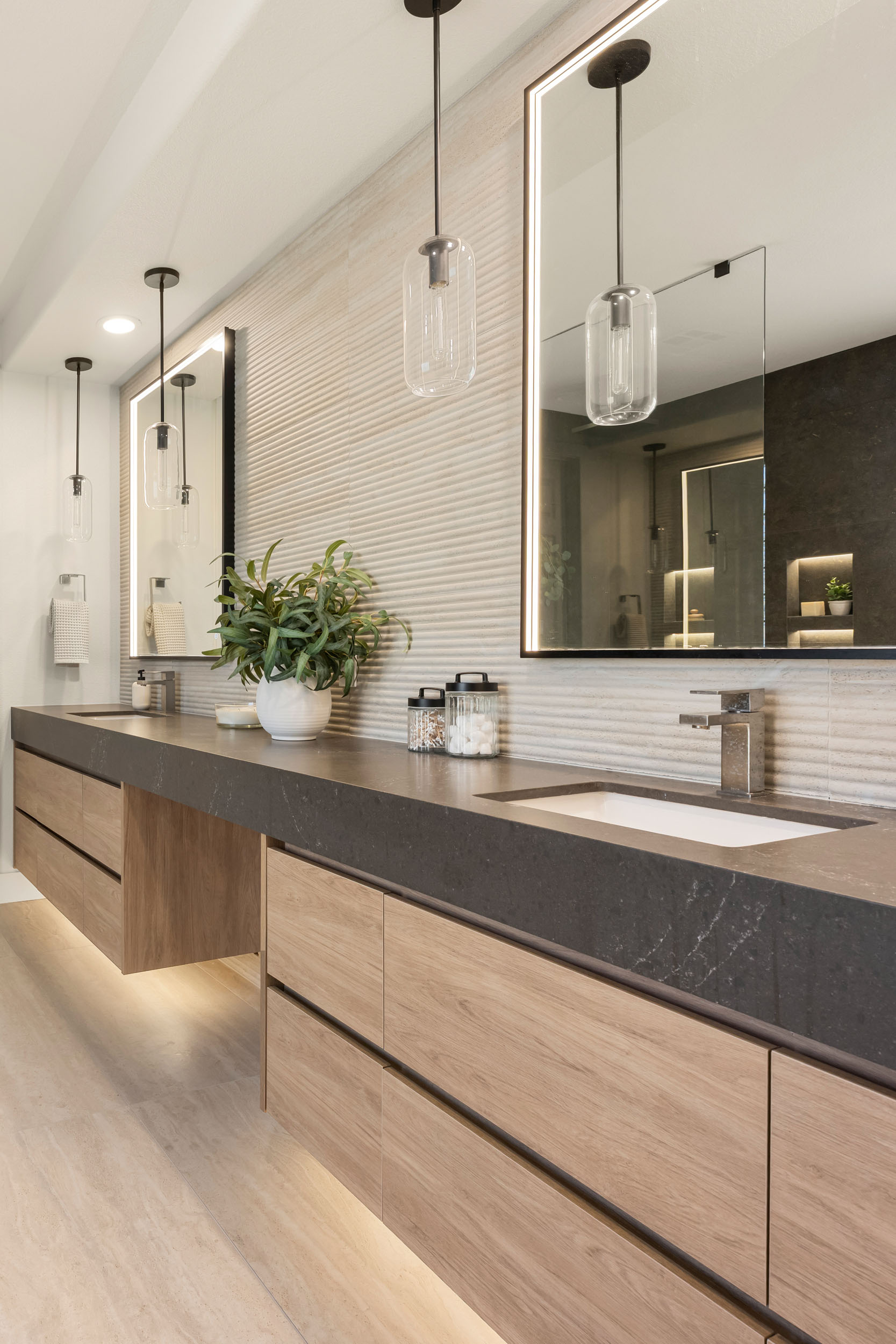 Primary bath vanity angle with long floating oak double vanity, glass cylinder pendants, black-framed mirrors, and ribbed tile backsplash in Summerlin remodel by Kingdom & Co.